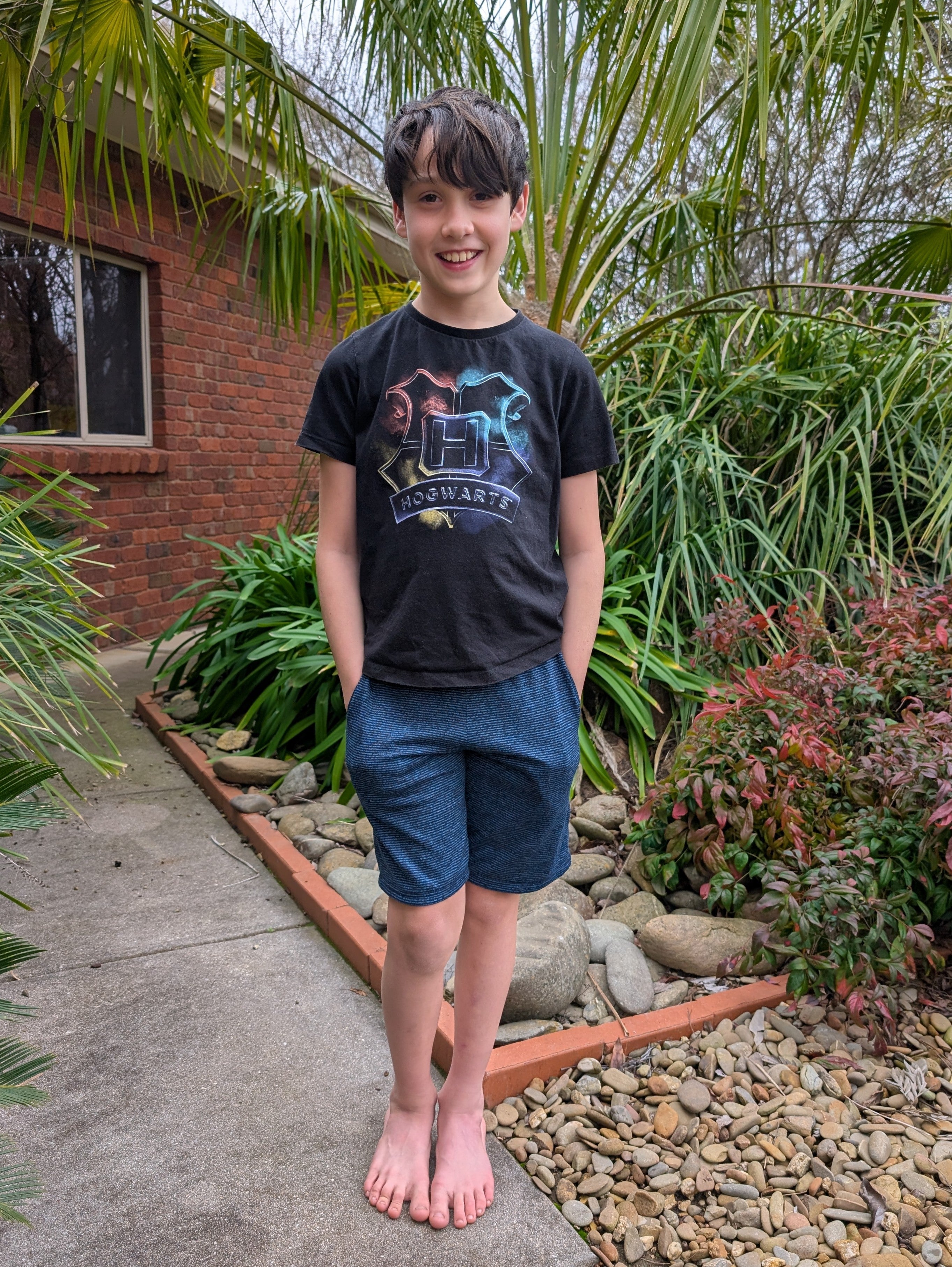 Young boy standing outdoors in a garden with plants and a house in the background wearing heathered athletic shorts with pockets made using the Caleb Athletic Sewing Pattern by Ellie and Mac Patterns.