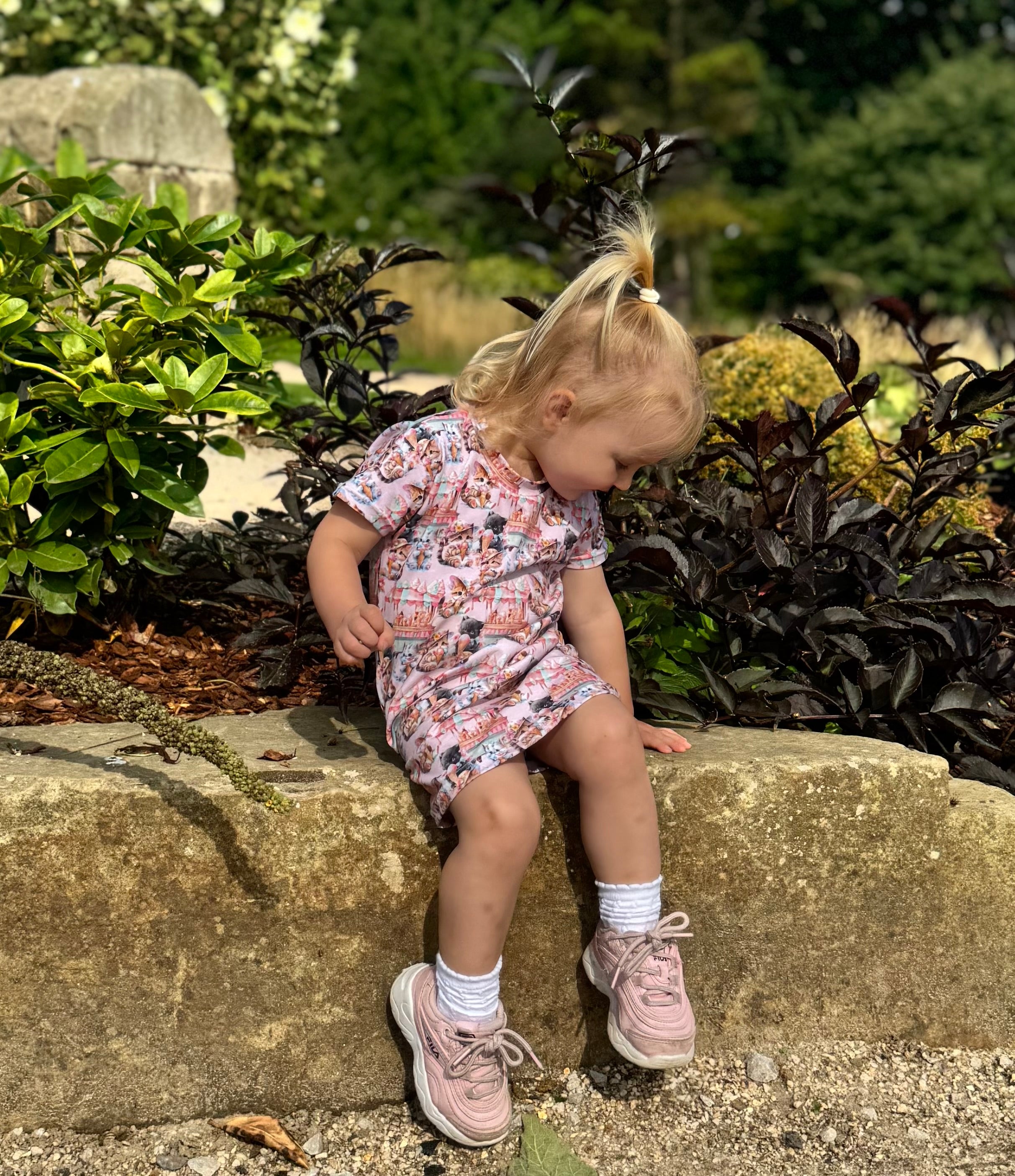 Young girl in a floral dress sitting on a stone ledge in a garden.