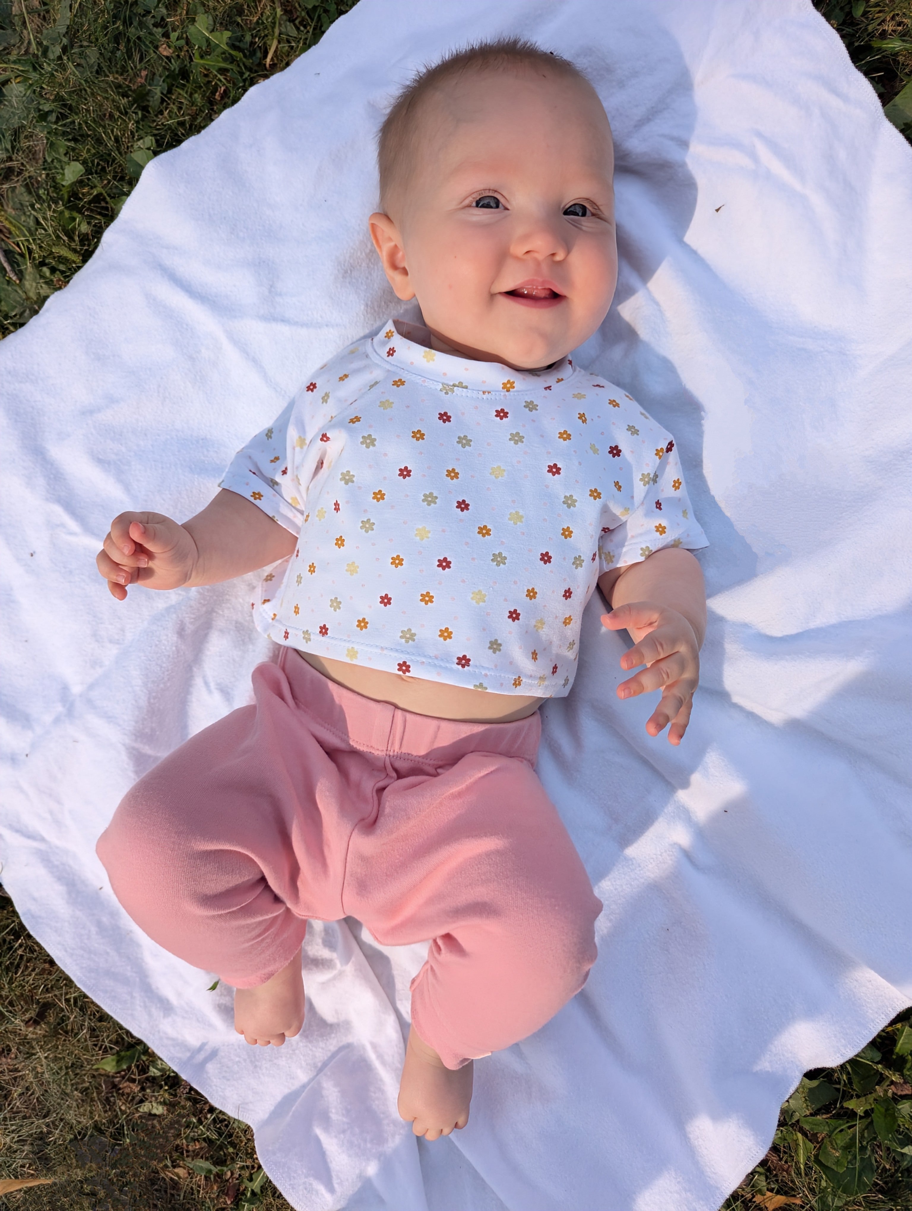 Baby wearing a white crop top with colorful patterns and pink pants, lying on a white blanket outdoors. Crop top made using the Kayla Sewing Pattern by Ellie and Mac.