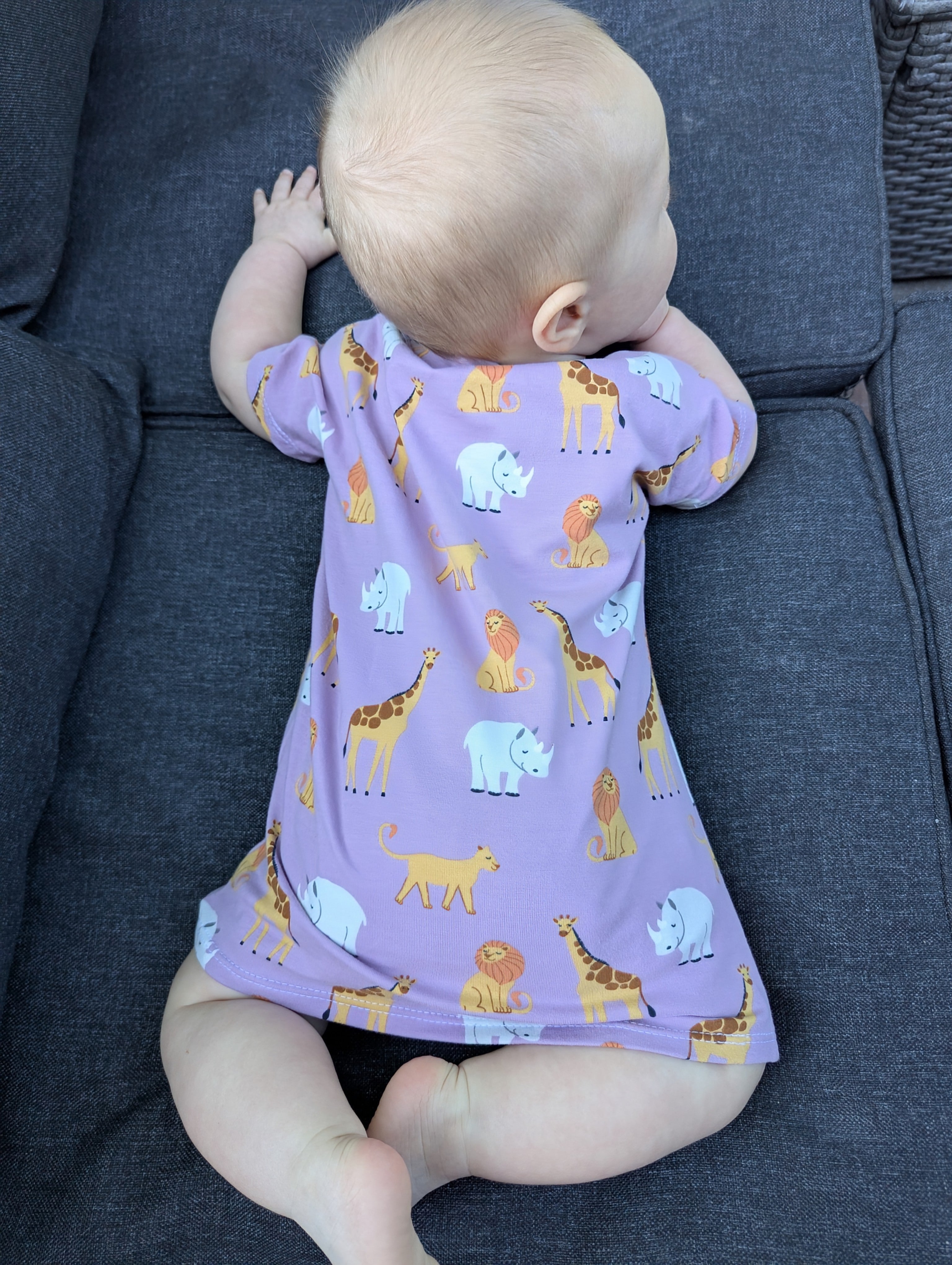 Baby wearing a purple dress with animal prints sitting on a dark blue couch.