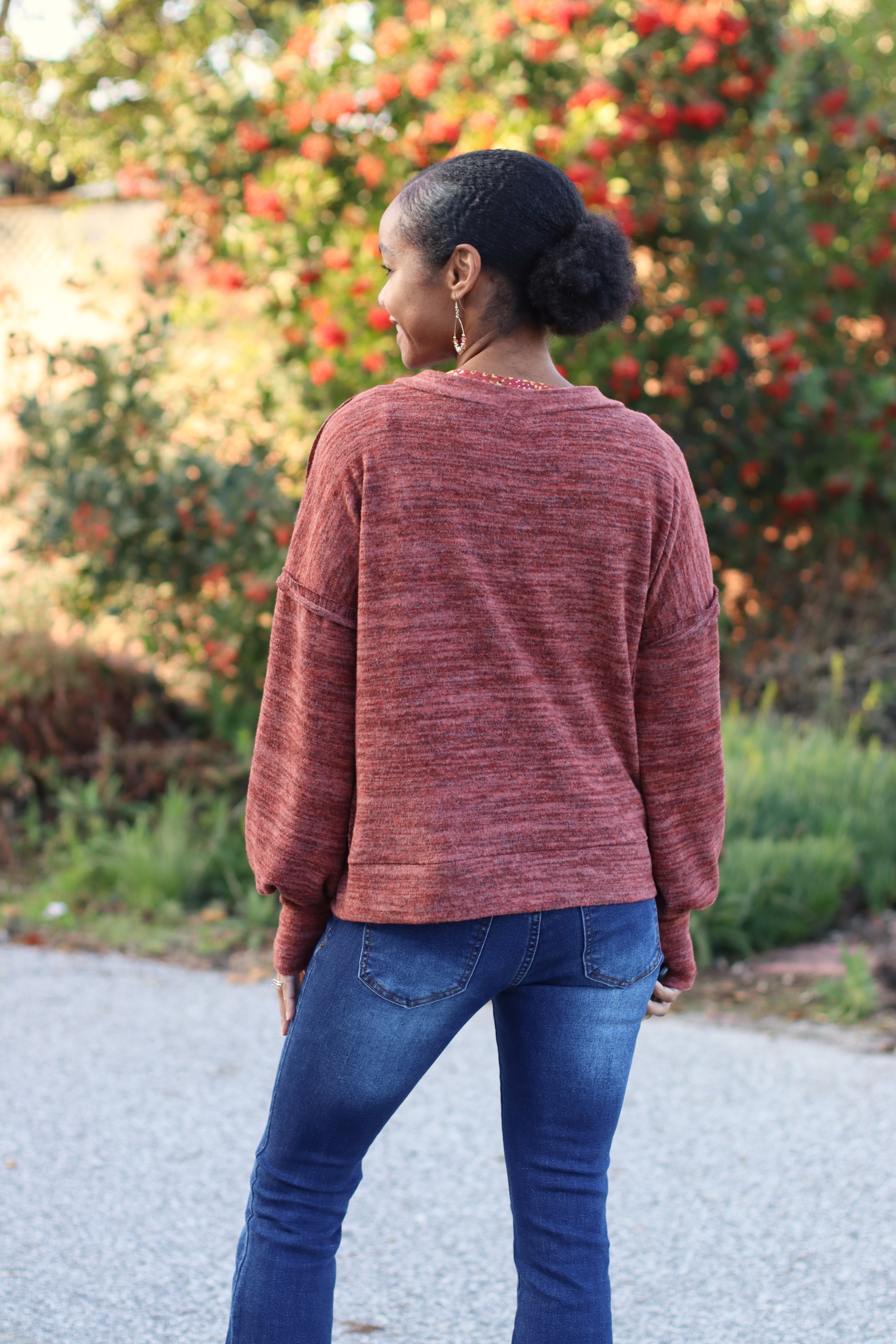 Person wearing a maroon sweater and blue jeans standing outdoors with greenery in the background