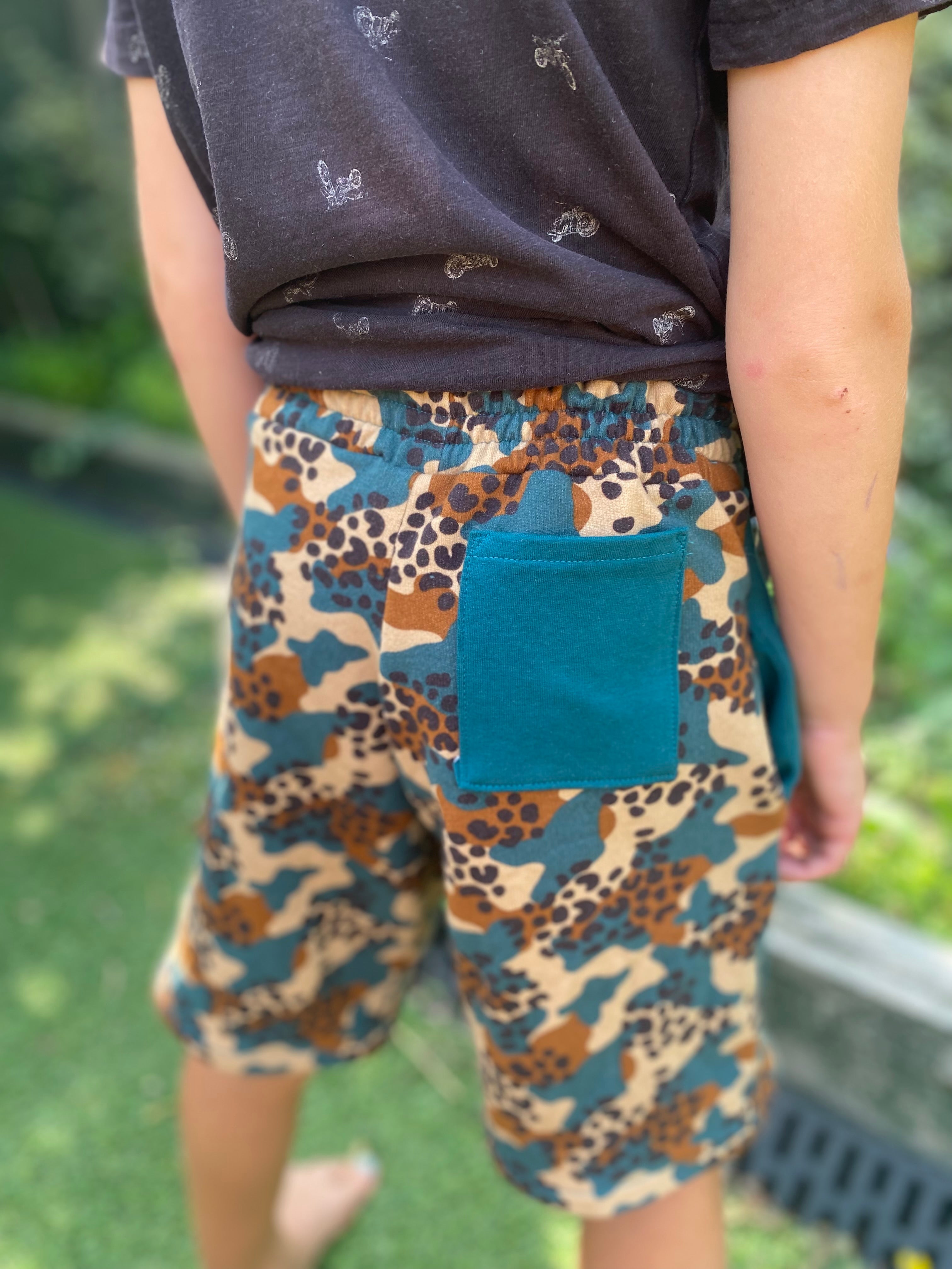 Person wearing camouflage shorts with a blue pocket in an outdoor setting made using the Caleb Athletic Sewing Pattern by Ellie and Mac Patterns.