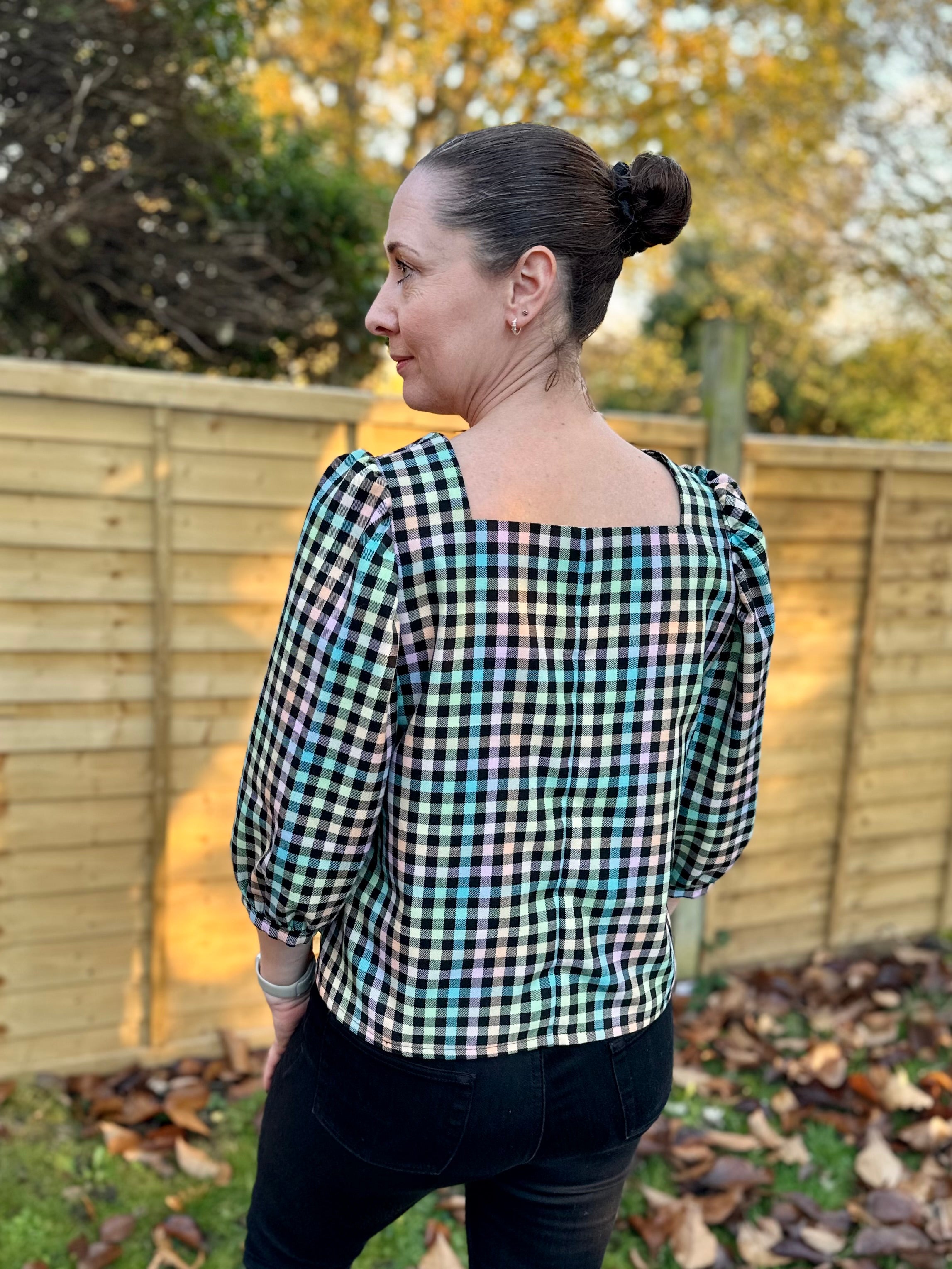 Woman wearing a black and white checkered handmade square neck blouse outdoors with trees and wooden fence in the background.