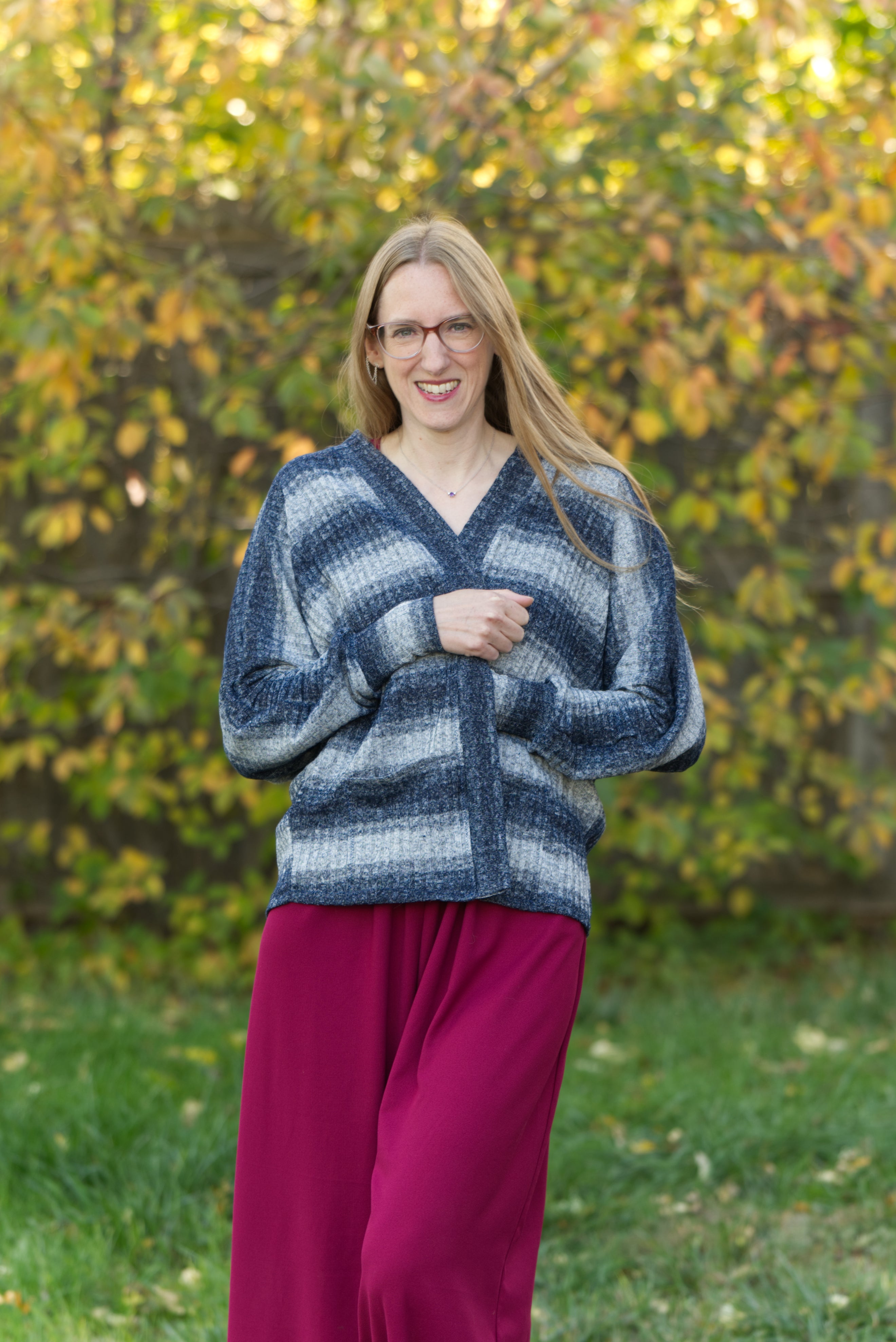 Woman wearing a blue striped sweater cardigan over a pink romper standing in front of green foliage. Cardigan made using the Ellie and Mac sewing pattern Celeste