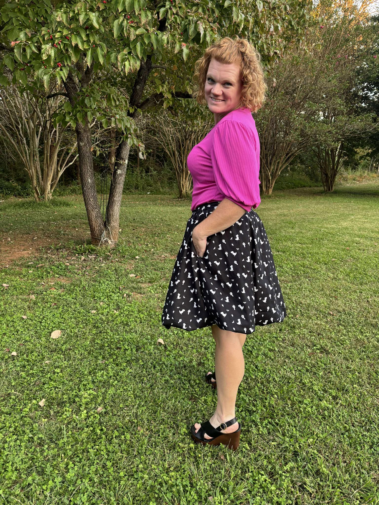 Woman in a hot pink top and black, knee-length bubble skirt, standing outdoors on grass with trees in the background.