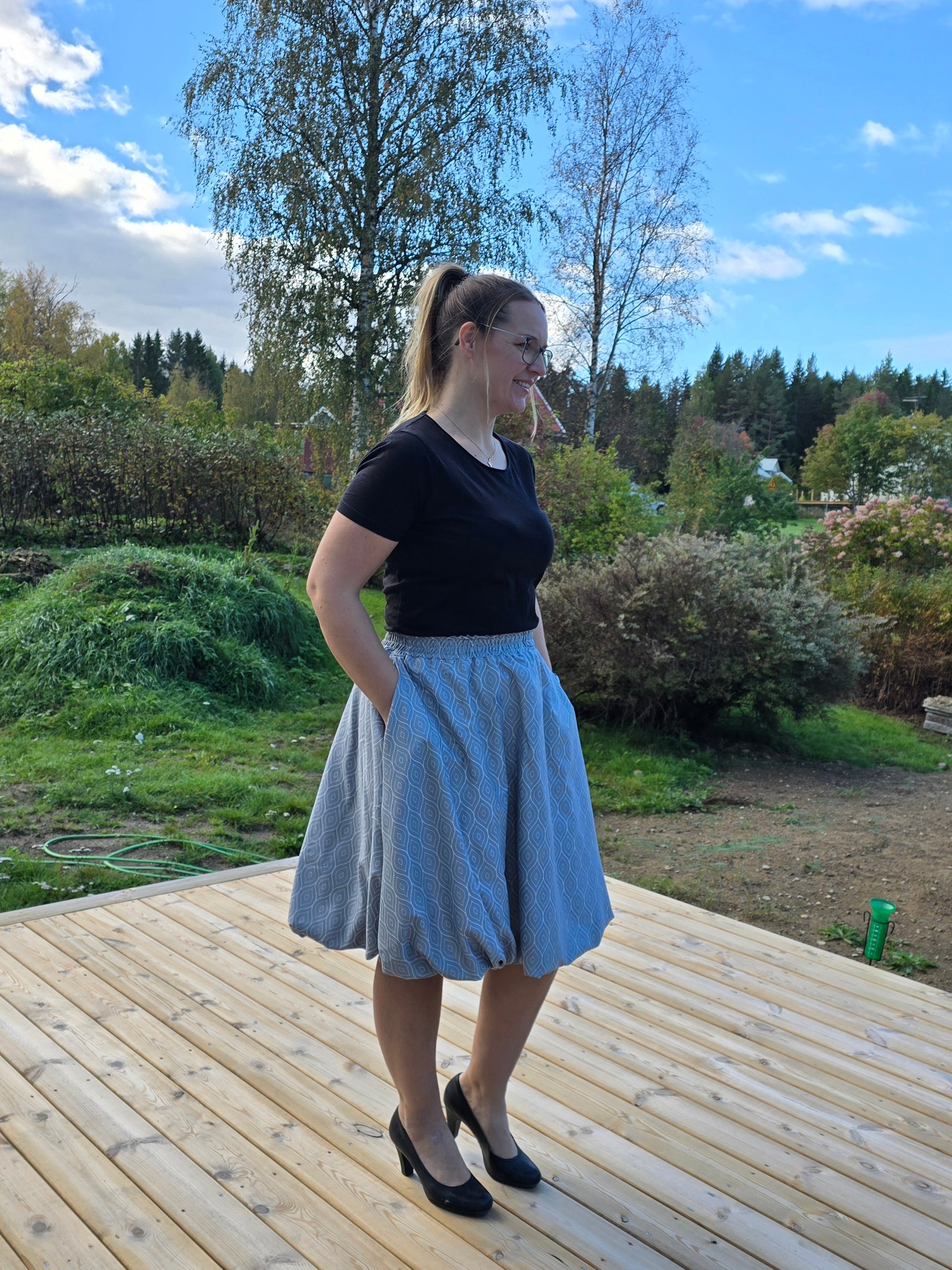 Woman standing on a wooden deck with trees and sky in the background. Wearing a knee-length bubble skirt with pocket and high heels.