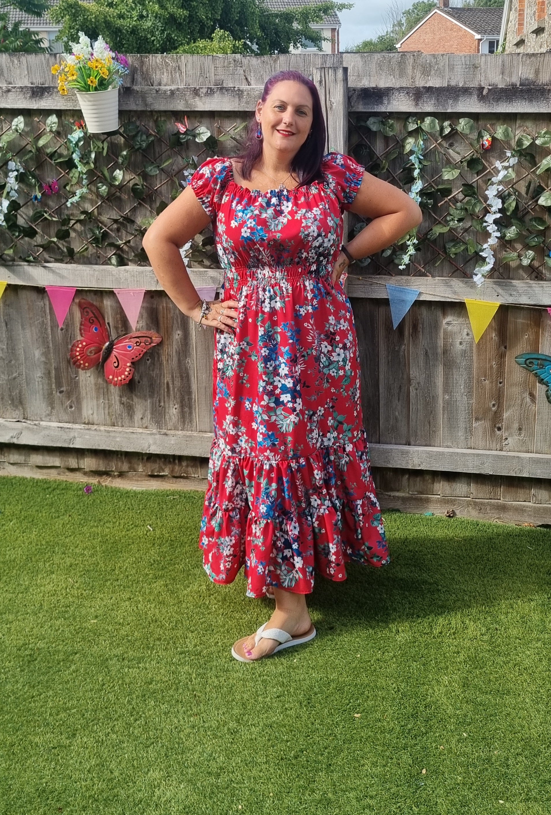 Woman in a red floral dress with cap sleeves and shirred waist, standing in a garden with a wooden fence and decorative flags in the background. Shantelle Shirred Dress sewing pattern used to make it.
