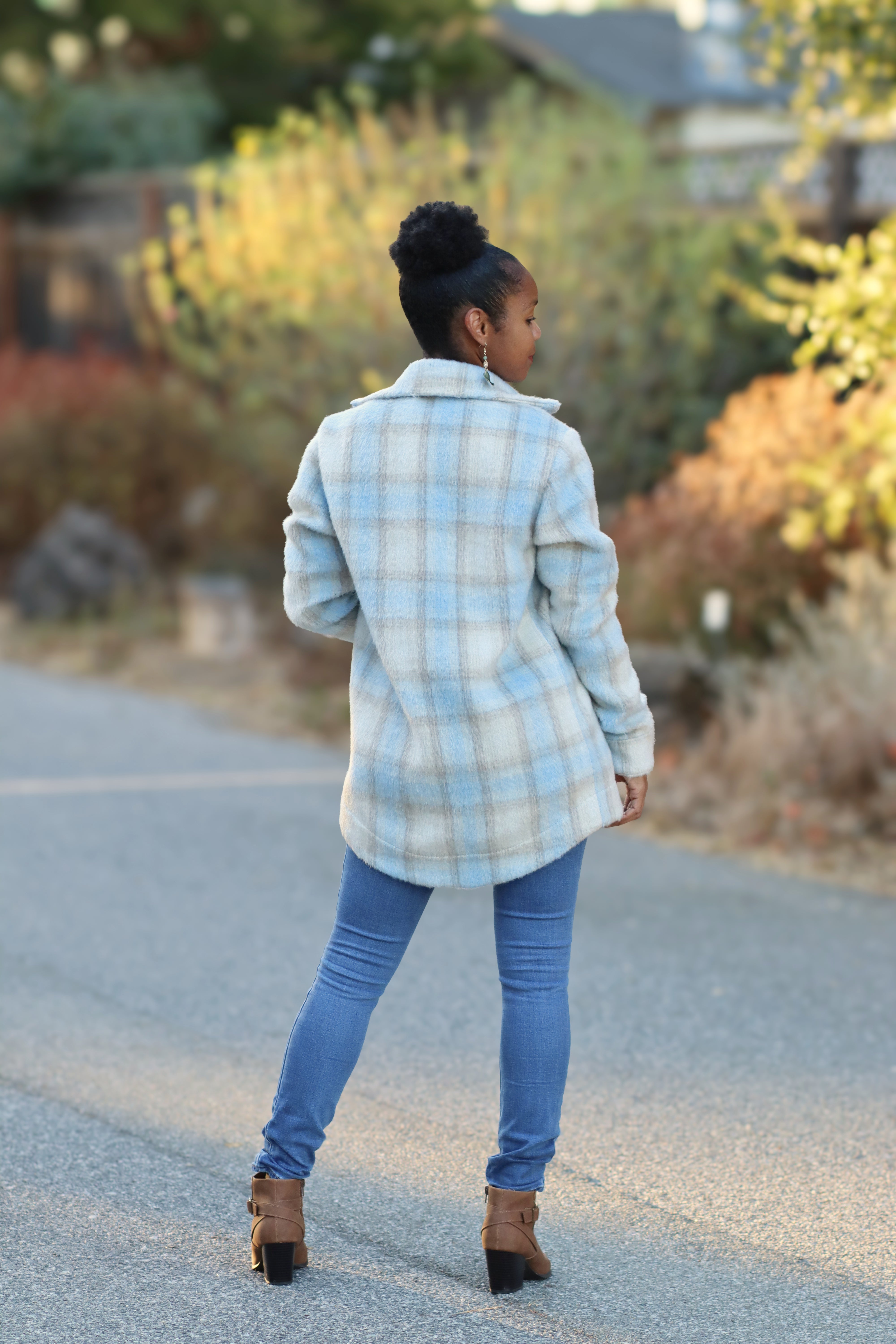 Back view of a woman outside in a street modeling a thick flannel shacket that is mid-thigh and has a curved hem. The flannel is blue and tan. She is wearing skinny jeans and boots.
