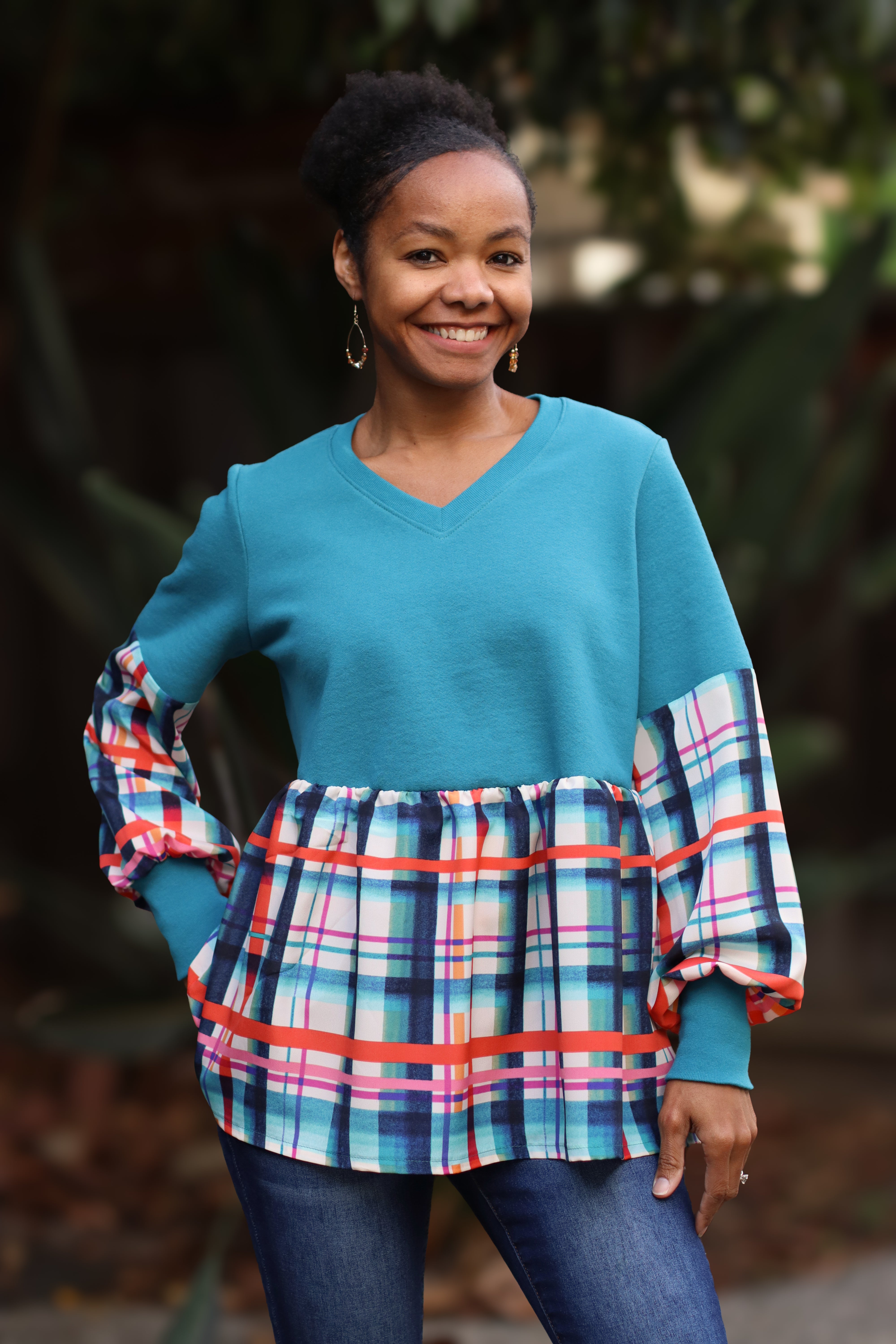 Woman wearing a turquoise top with plaid gathered skirt and bishop sleeve details against a blurred natural background.