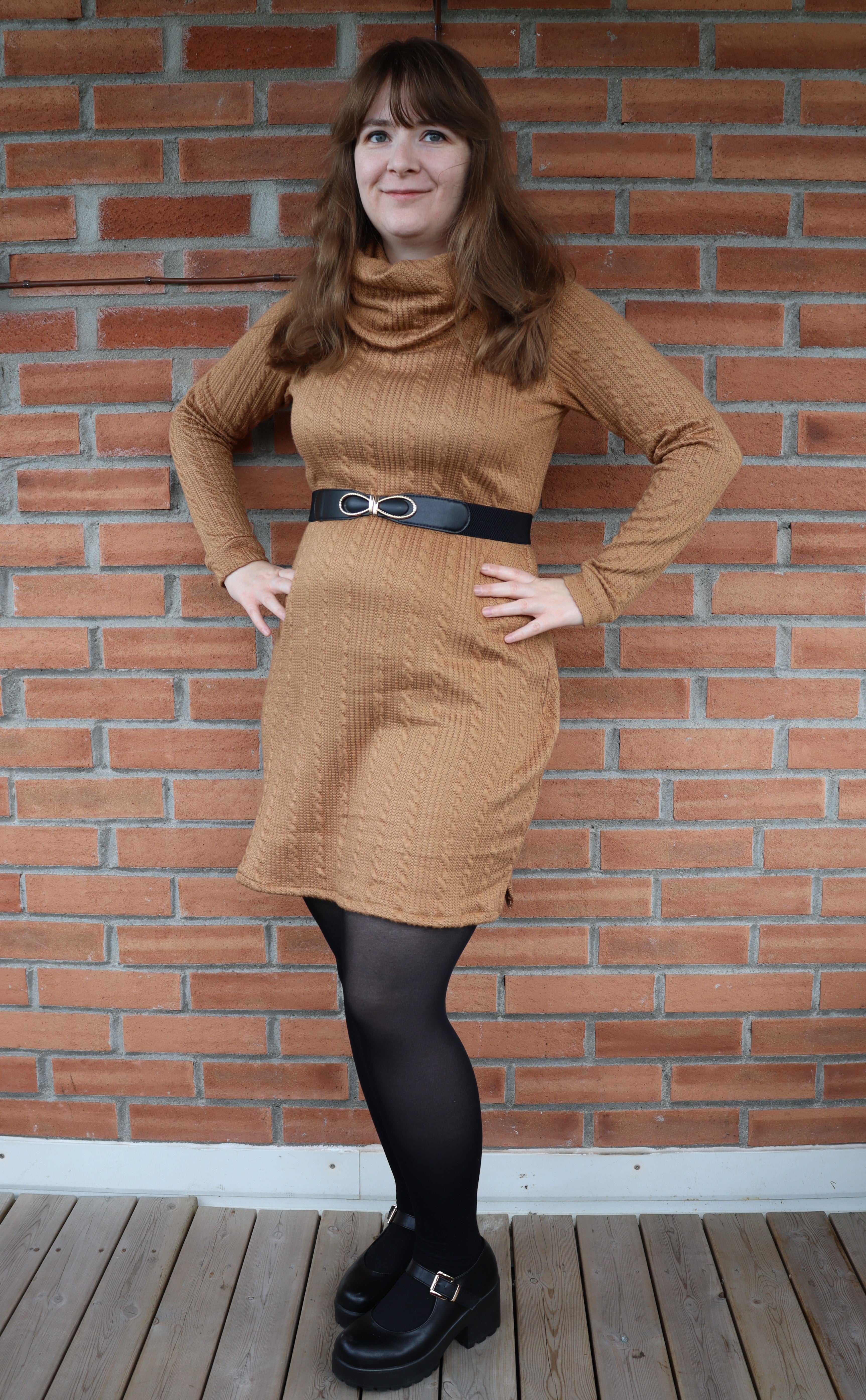 Woman wearing a brown dress with a belt against a brick wall.