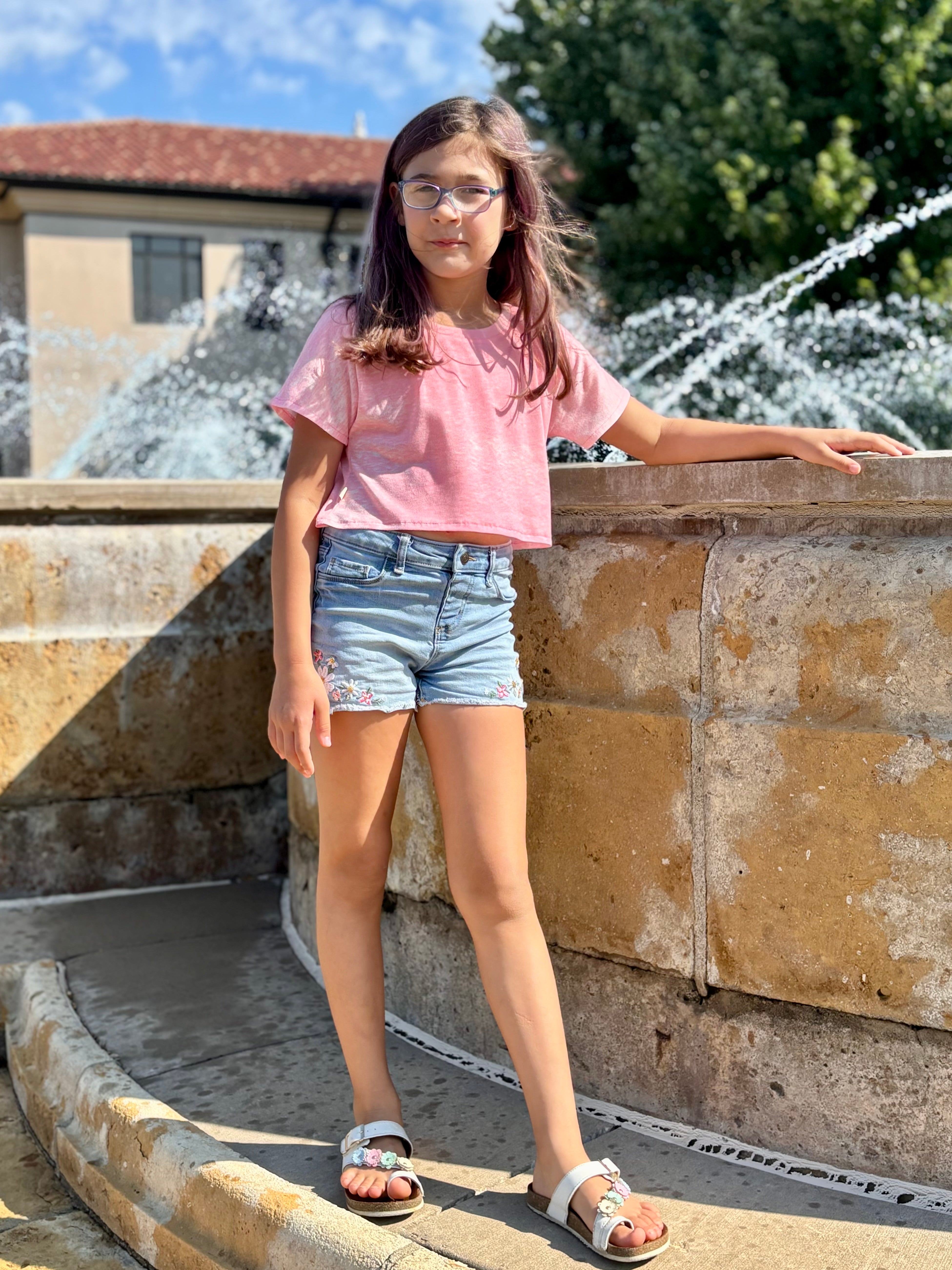 Young girl in a pink shirt and denim shorts standing near a fountain.
