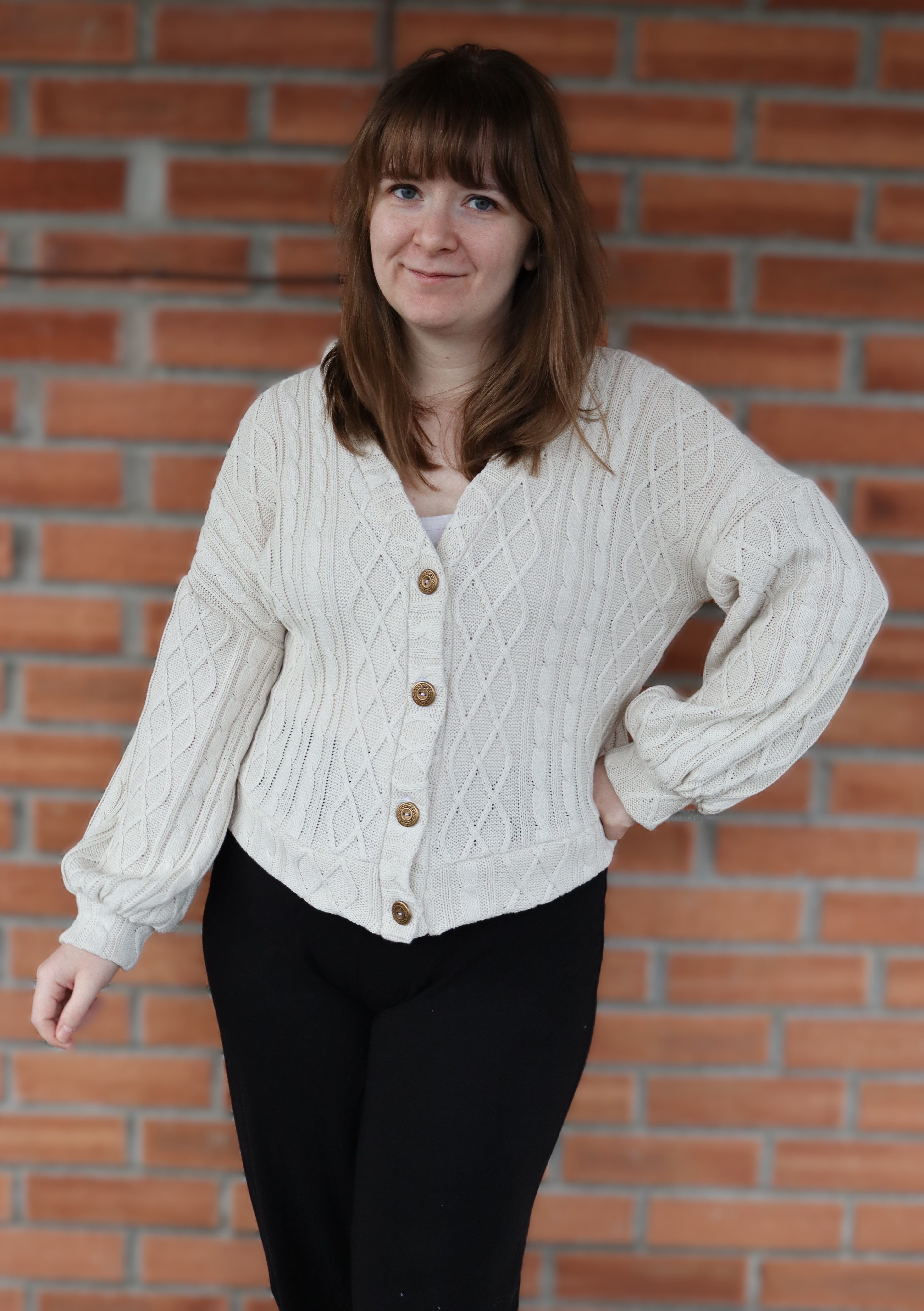 Woman wearing a white cable knit cardigan with wooden buttons against a brick wall.