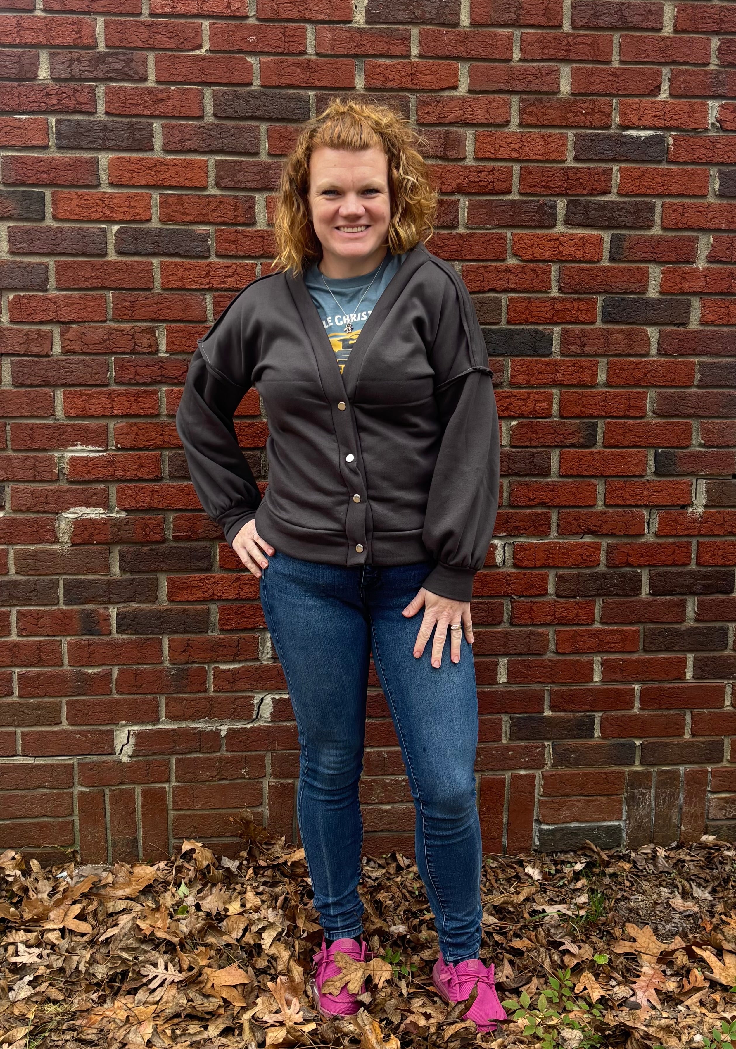 Person wearing a dark gray cardigan with bishop sleeves over a graphic t-shirt, standing against a brick wall with fallen leaves on the ground.