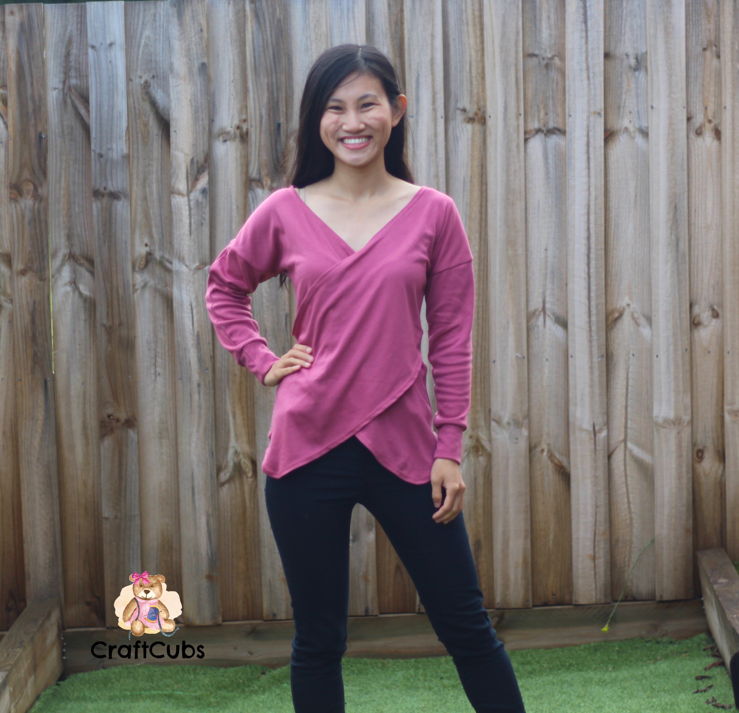 Woman wearing a pink long-sleeve dolman top and black pants standing in front of a wooden fence.
