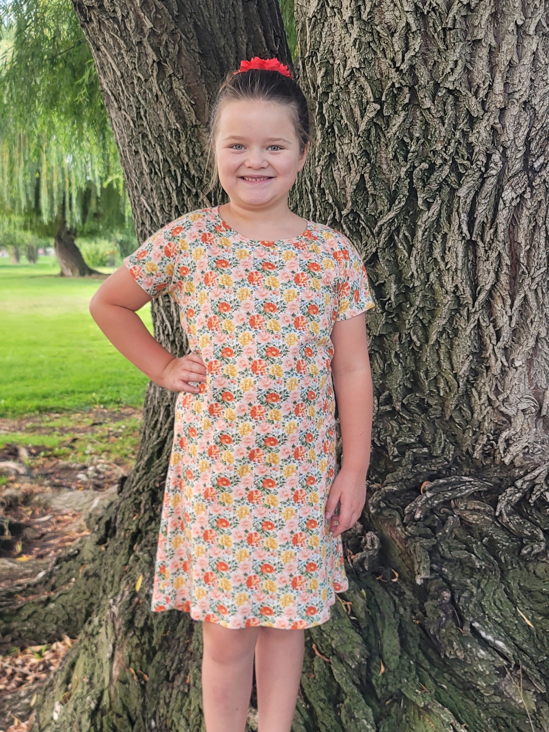 Child in a floral dress standing between two large trees with a park background. dolman dress sewing pattern.
