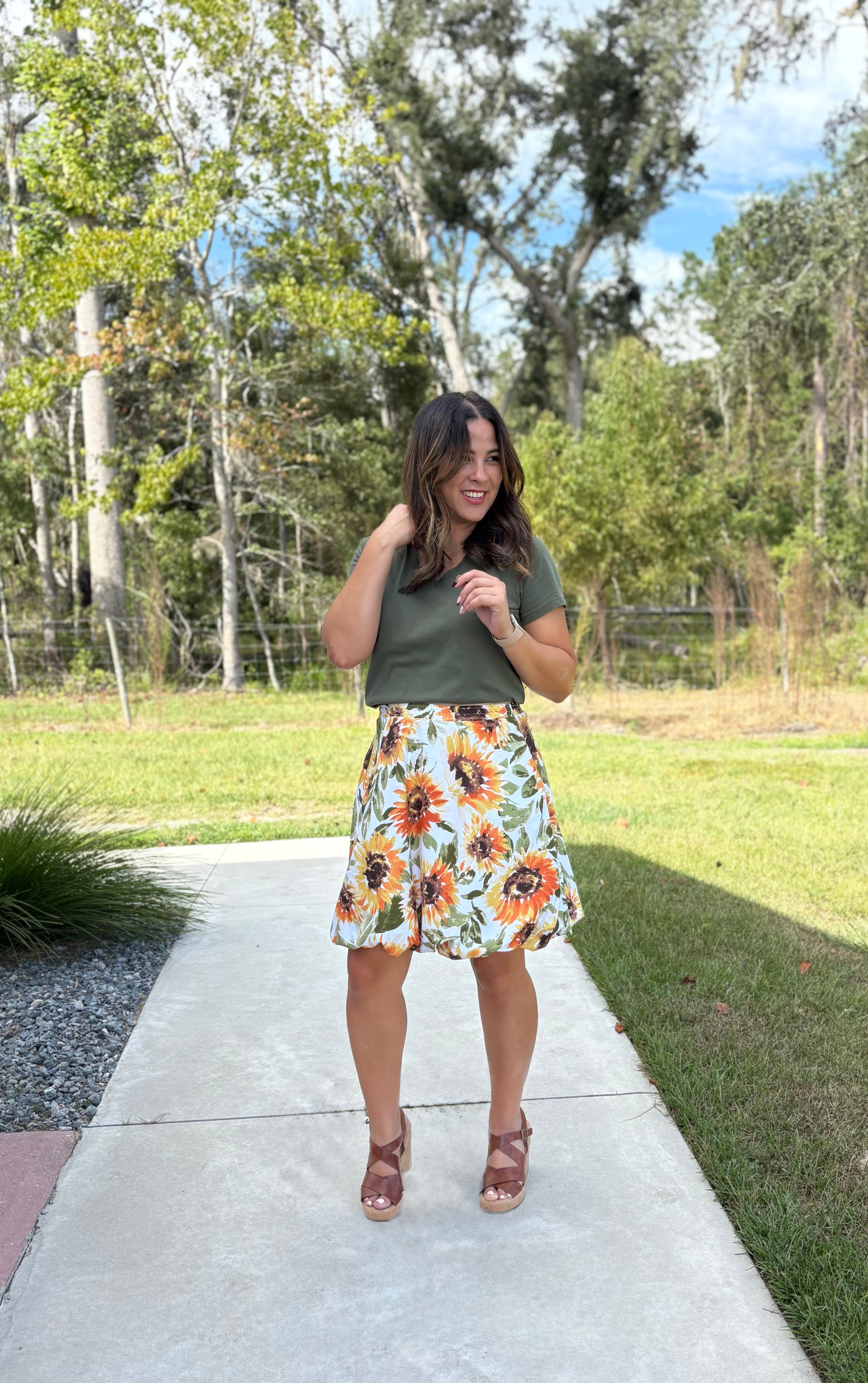 Woman in a green top and sunflower knee-length bubble skirt standing on a sidewalk with trees in the background. Perfect fall skirt.