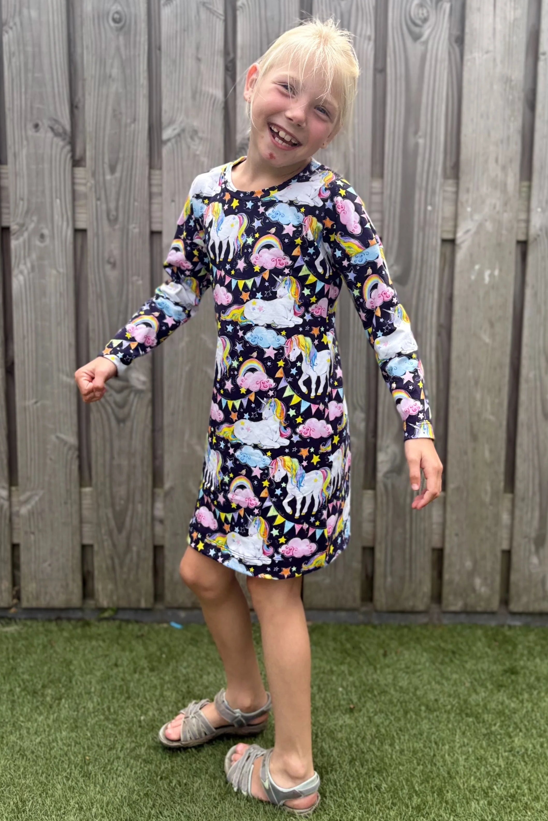 Child wearing a colorful dress with animal pattern standing in front of a wooden fence.