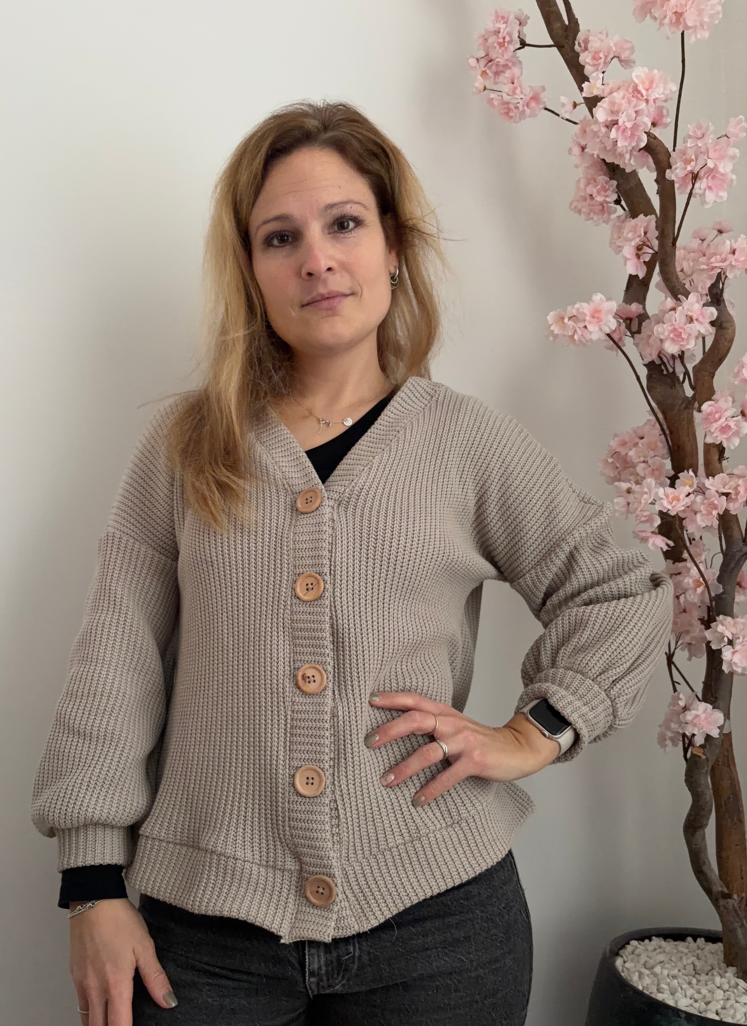 Woman wearing a beige cardigan standing next to a decorative plant with pink flowers.