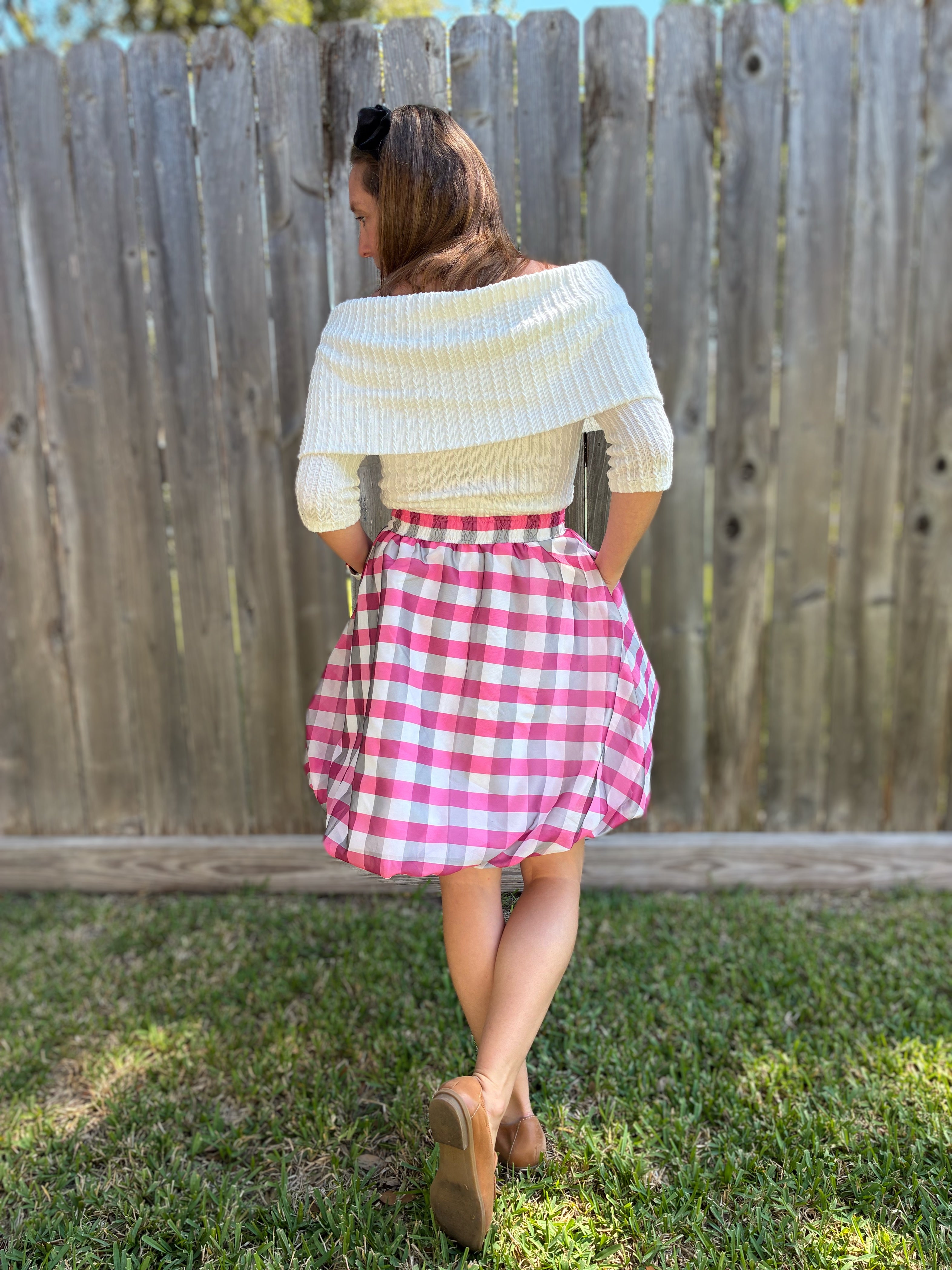 Person wearing a white sweater and pink checkered skirt standing in front of a wooden fence.