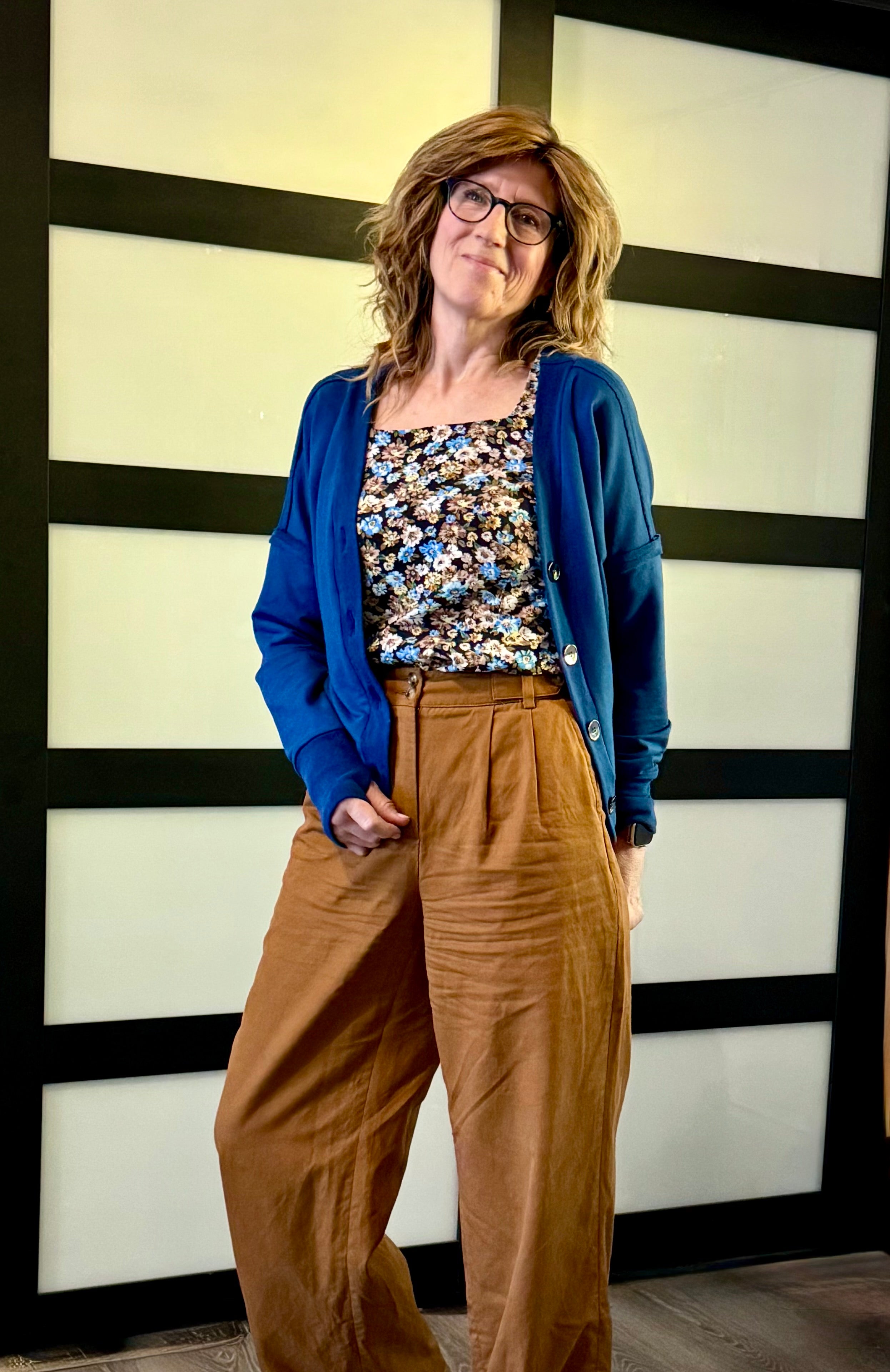 Woman wearing a blue cardigan, floral top, and brown pants standing against a black and white striped wall. Top and cardigan sewn using Ellie and Mac Sewing patterns