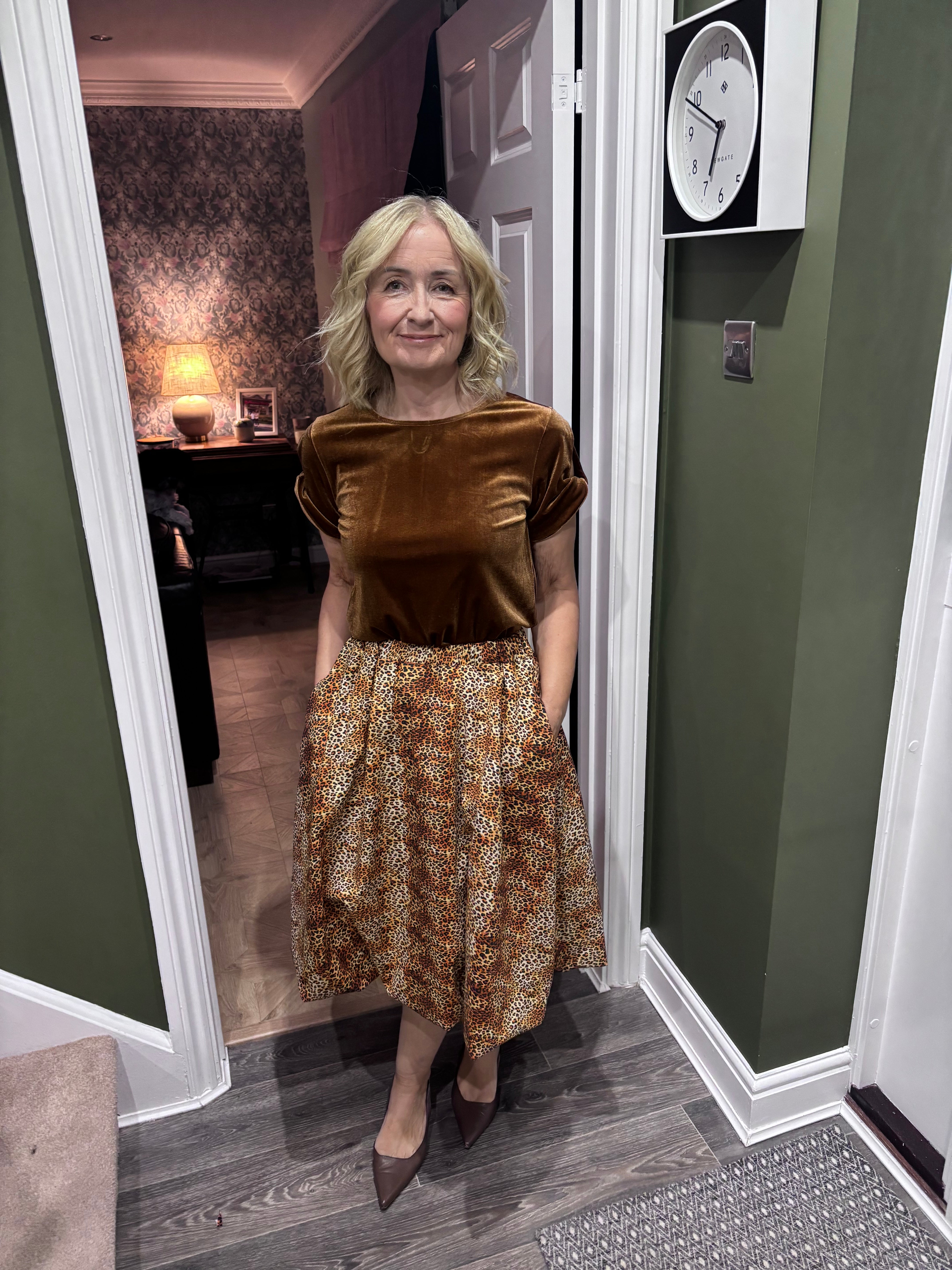 Woman in a sophisticated gold velvet top and midi bubble skirt, standing in a room with a clock on the wall.