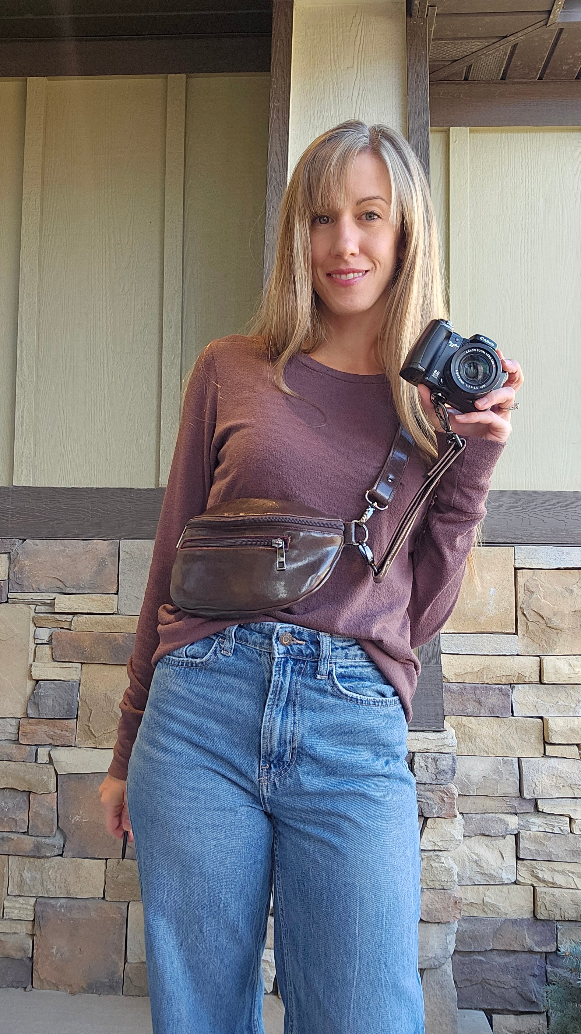 Woman in mauve long sleeve shirt and jeans, holding a camera that is attached to the camera strap that is an accessory to a brown leather belt bag the woman is wearing. Make your own belt bag with Ellie and Mac sewing patterns.