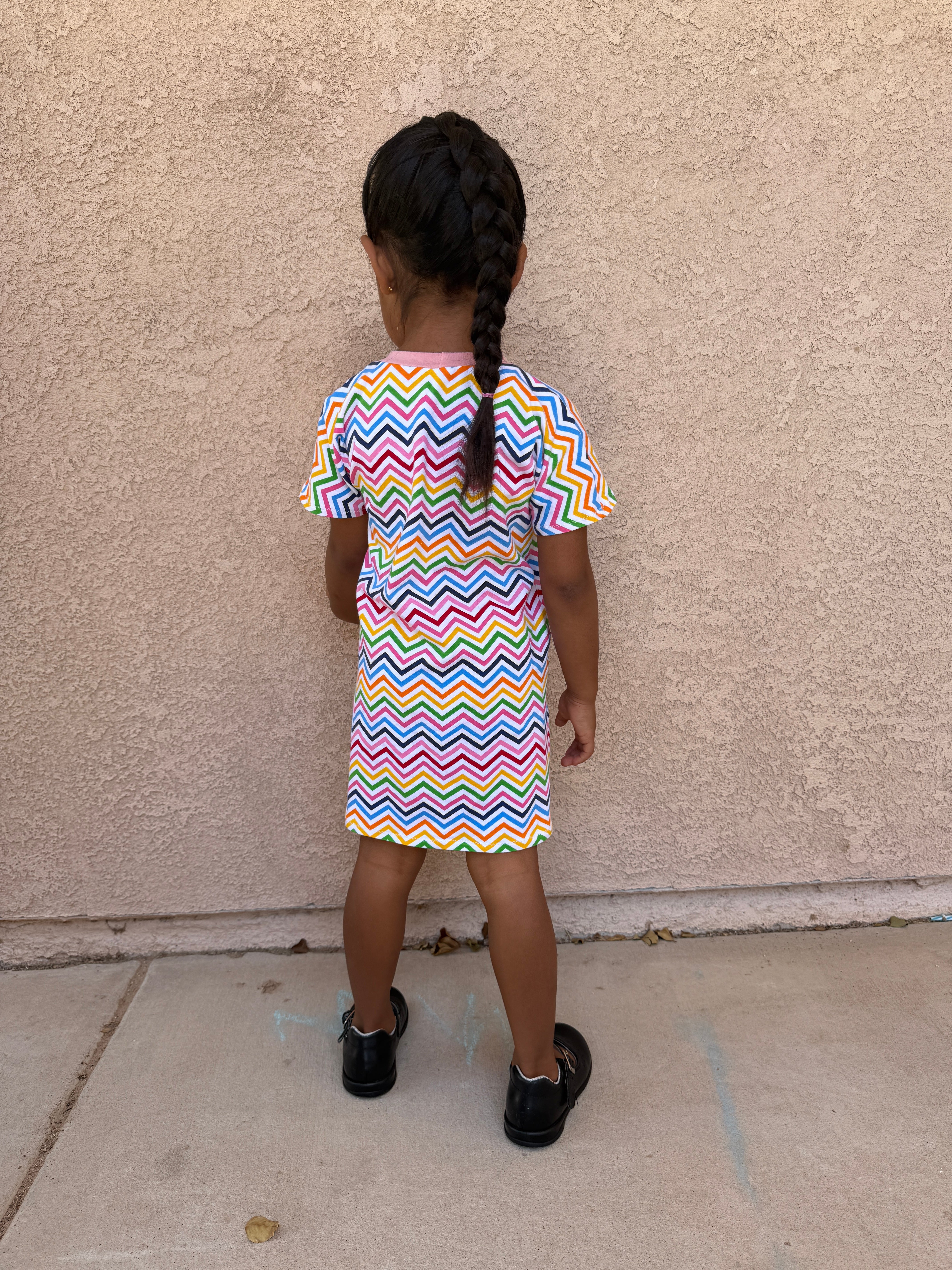 Child wearing a colorful zigzag dress standing on a sidewalk.