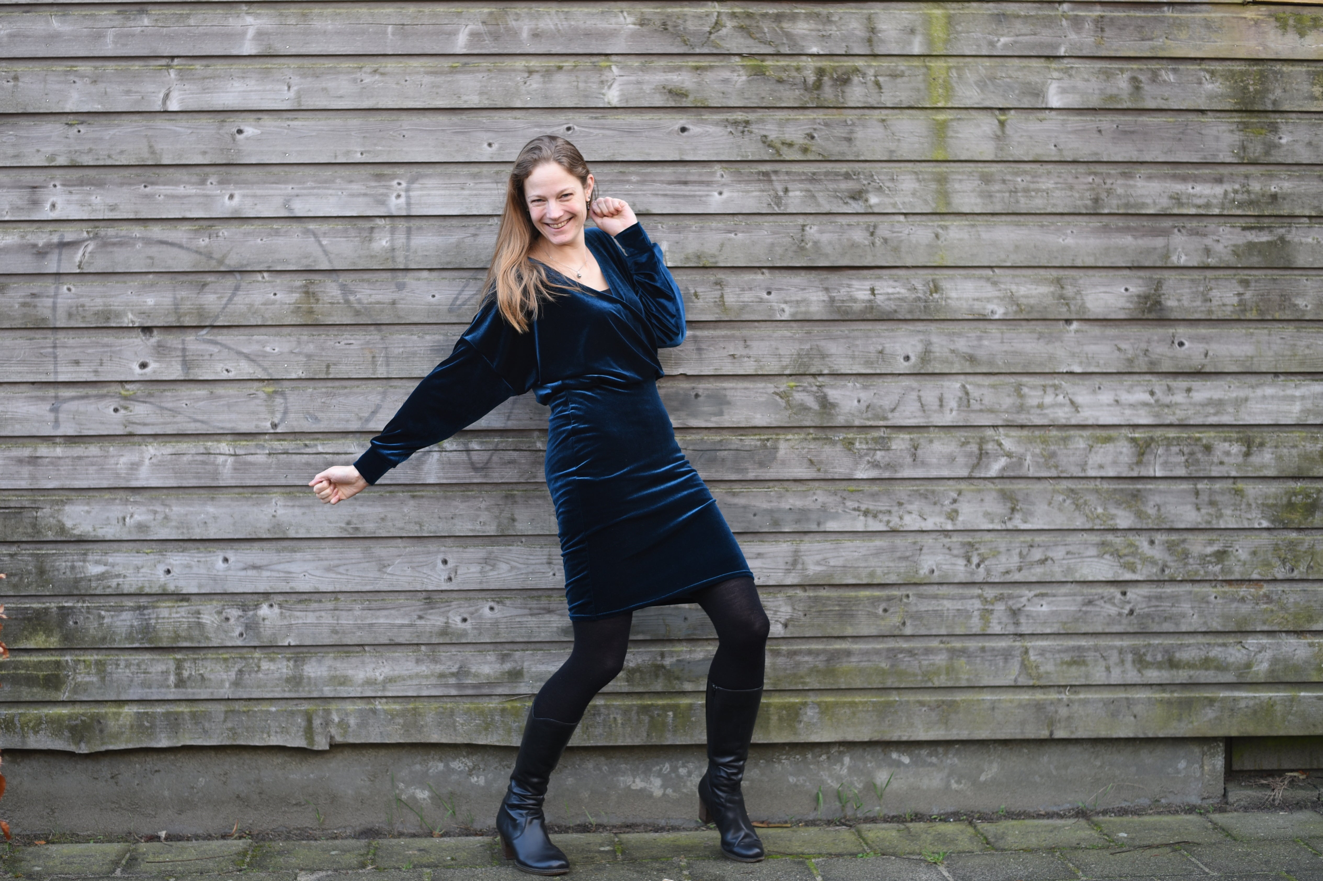 Woman in a blue velvet crossover wrap dress posing against a wooden wall with a big smile on her face.