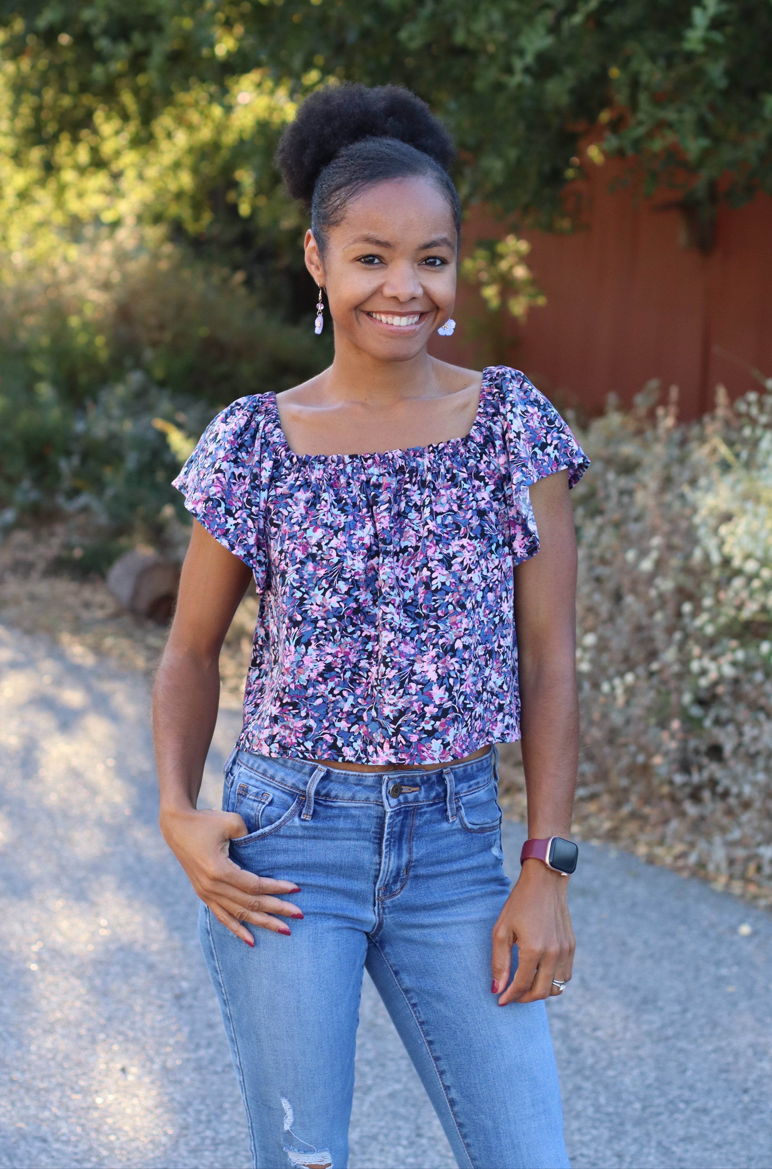 Woman wearing a floral top with flowy sleeves and jeans standing outdoors with greenery in the background. The top is made using the woven sewing pattern, Shantelle Shirred Blouse Pattern.