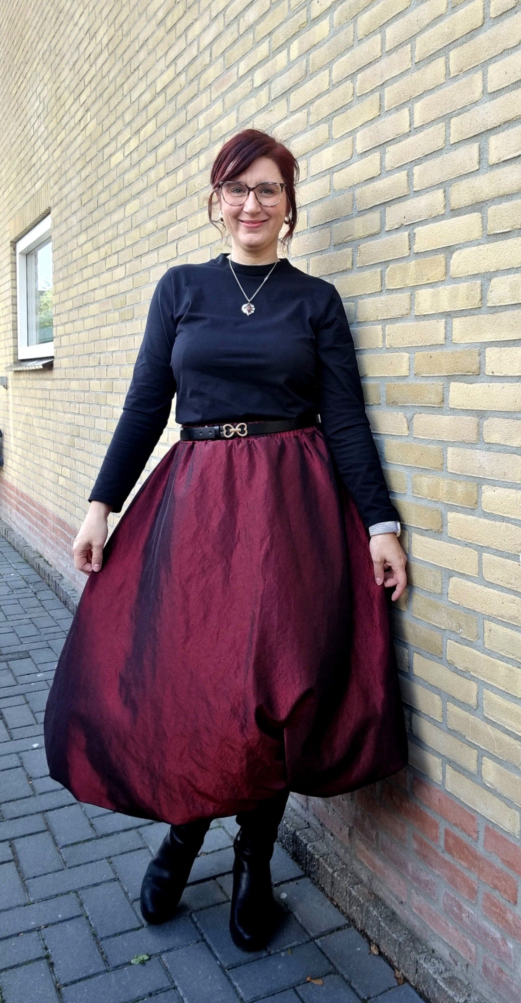 Person wearing a black top and burgundy midi bubble skirt standing against a brick wall. The skirt is made using taffeta fabric and Ellie and Mac's Sadie Bubble Skirt Pattern.