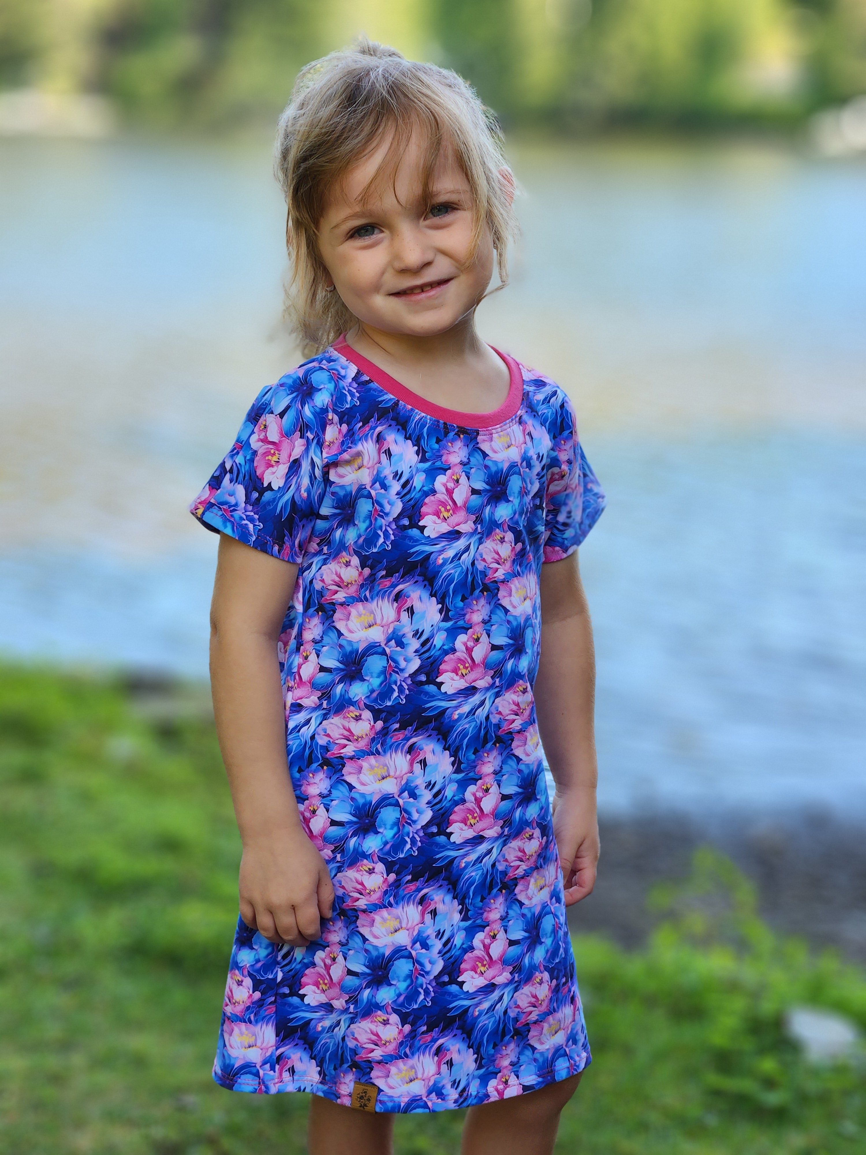 Child wearing a blue floral dress standing outdoors with a blurred natural background. Dress made using the Kayla Dolman Pattern by Ellie and Mac.