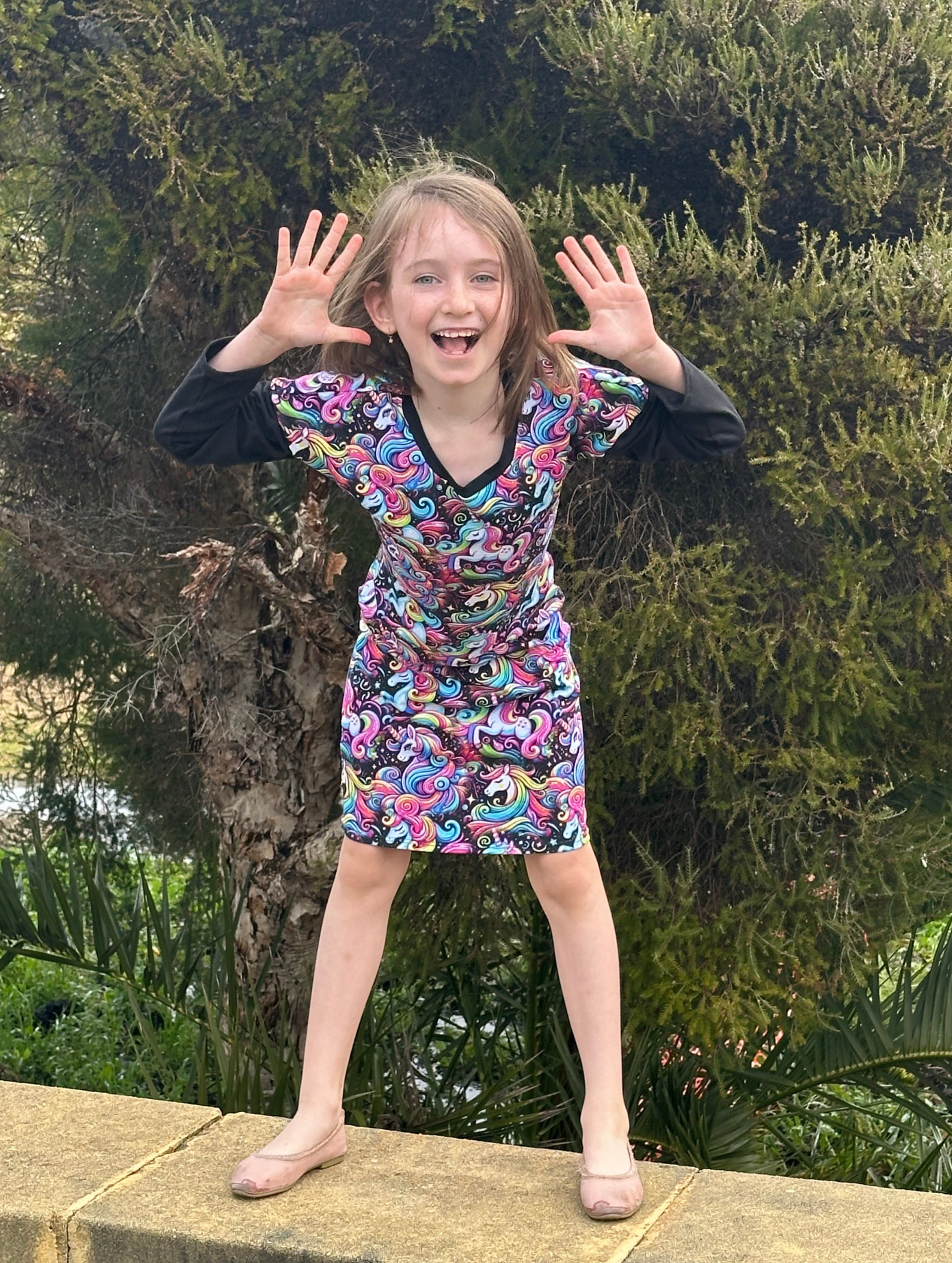 Child in a colorful dress standing on a stone ledge with greenery in the background.