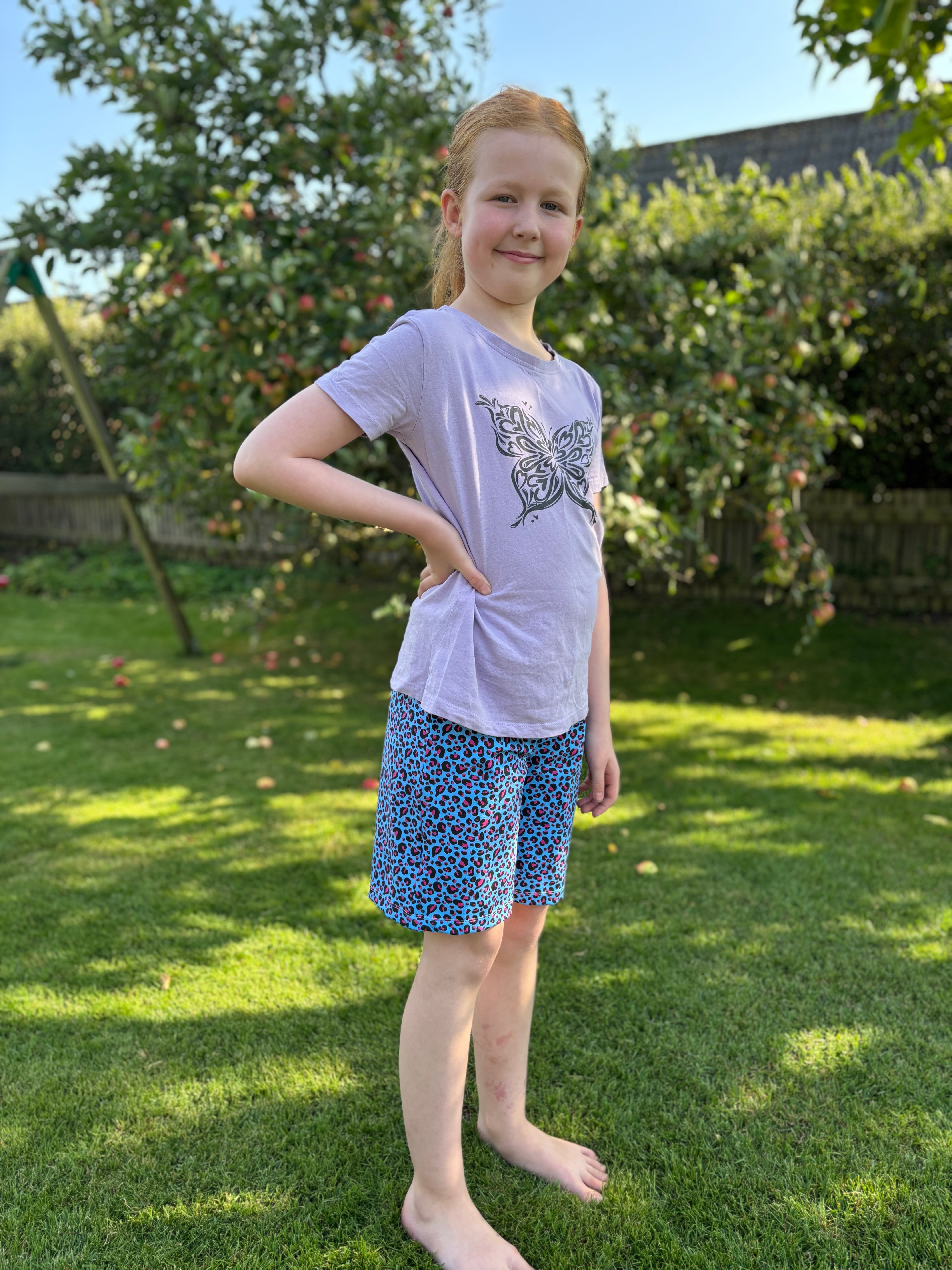Young girl standing on grass wearing a light purple t-shirt with a butterfly design and blue patterned shorts made using the Caleb Athletic Sewing Pattern by Ellie and Mac Patterns.