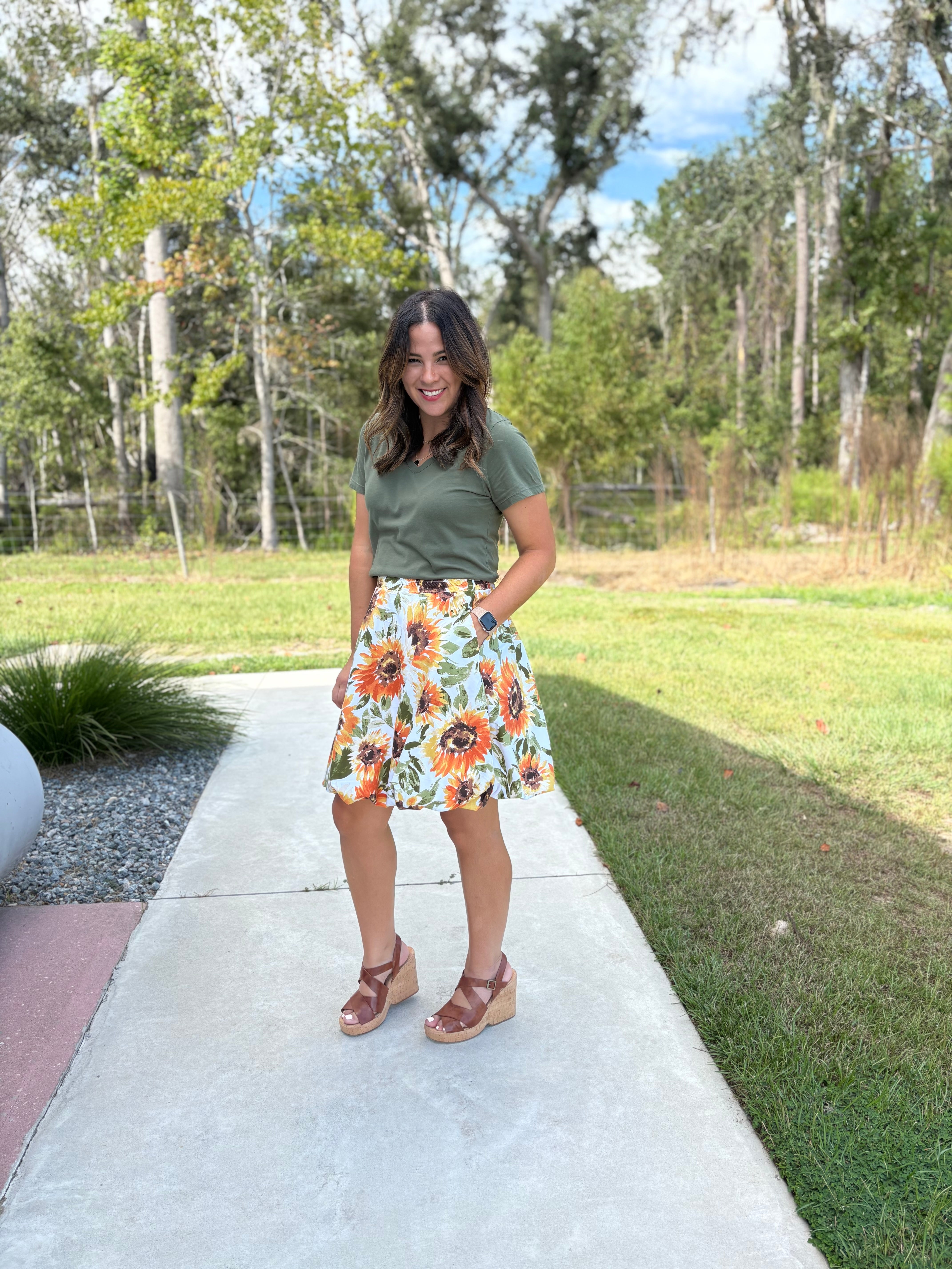 Woman standing on a sidewalk wearing a handmade green t-shirt and floral bubble skirt with trees in the background. The skirt is easy to make using Ellie and Mac's PDF sewing pattern.