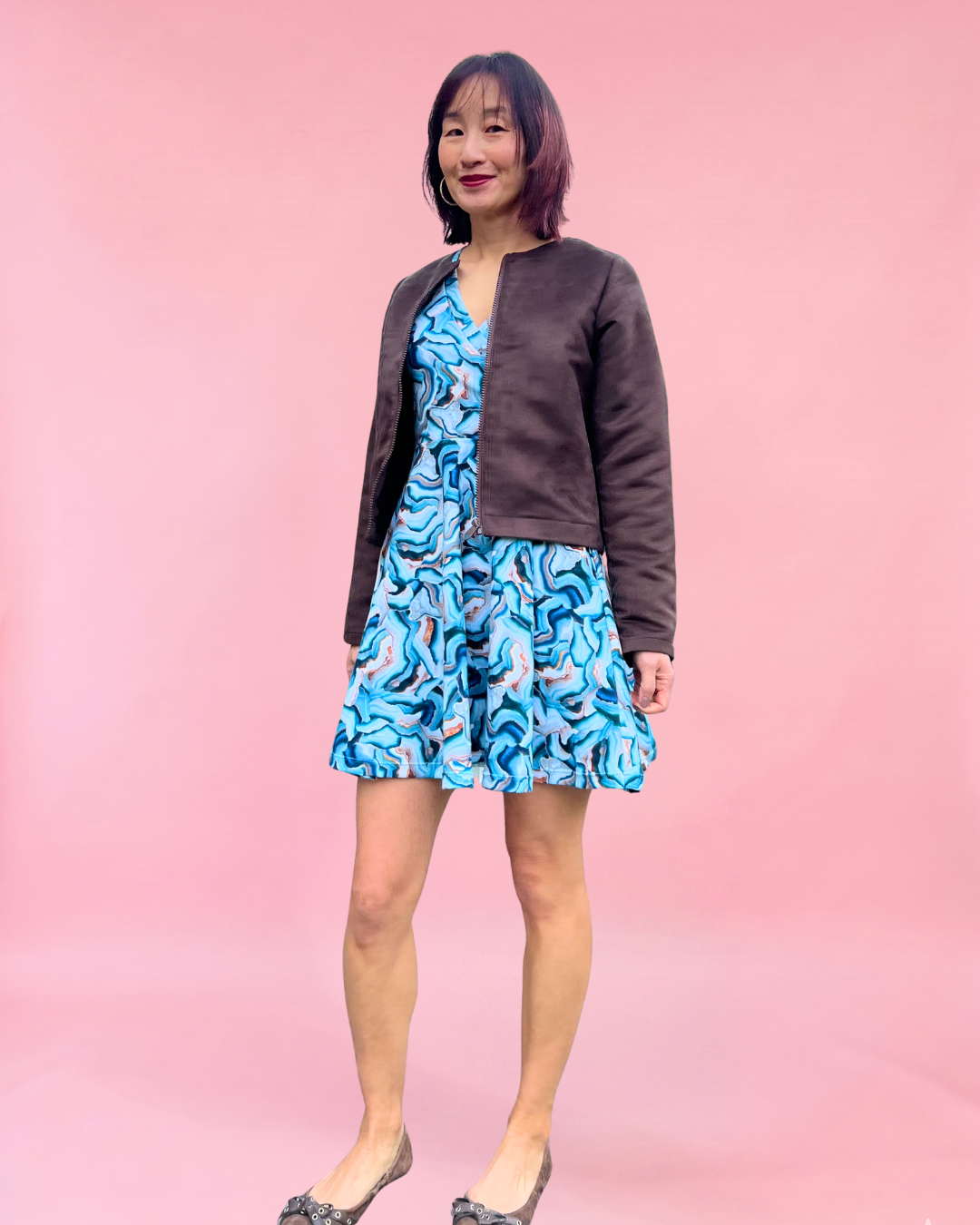 Woman wearing a blue patterned dress with a brown suede jacket against a pink background. Easy sewing patterns for beginners.