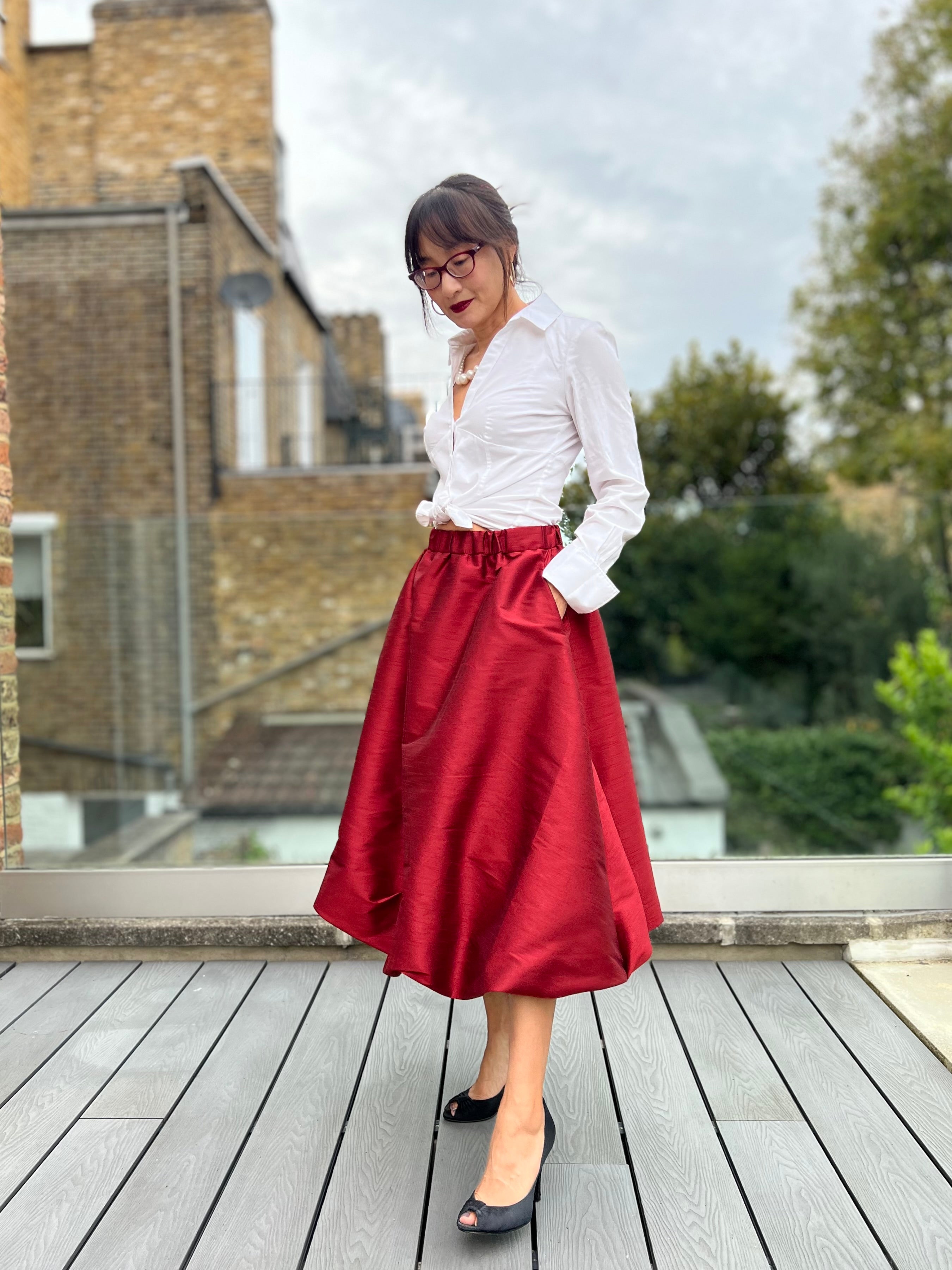 Woman wearing a white blouse and red taffeta bubble midi skirt on a wooden deck with a building and trees in the background. Easy PDF skirt pattern.