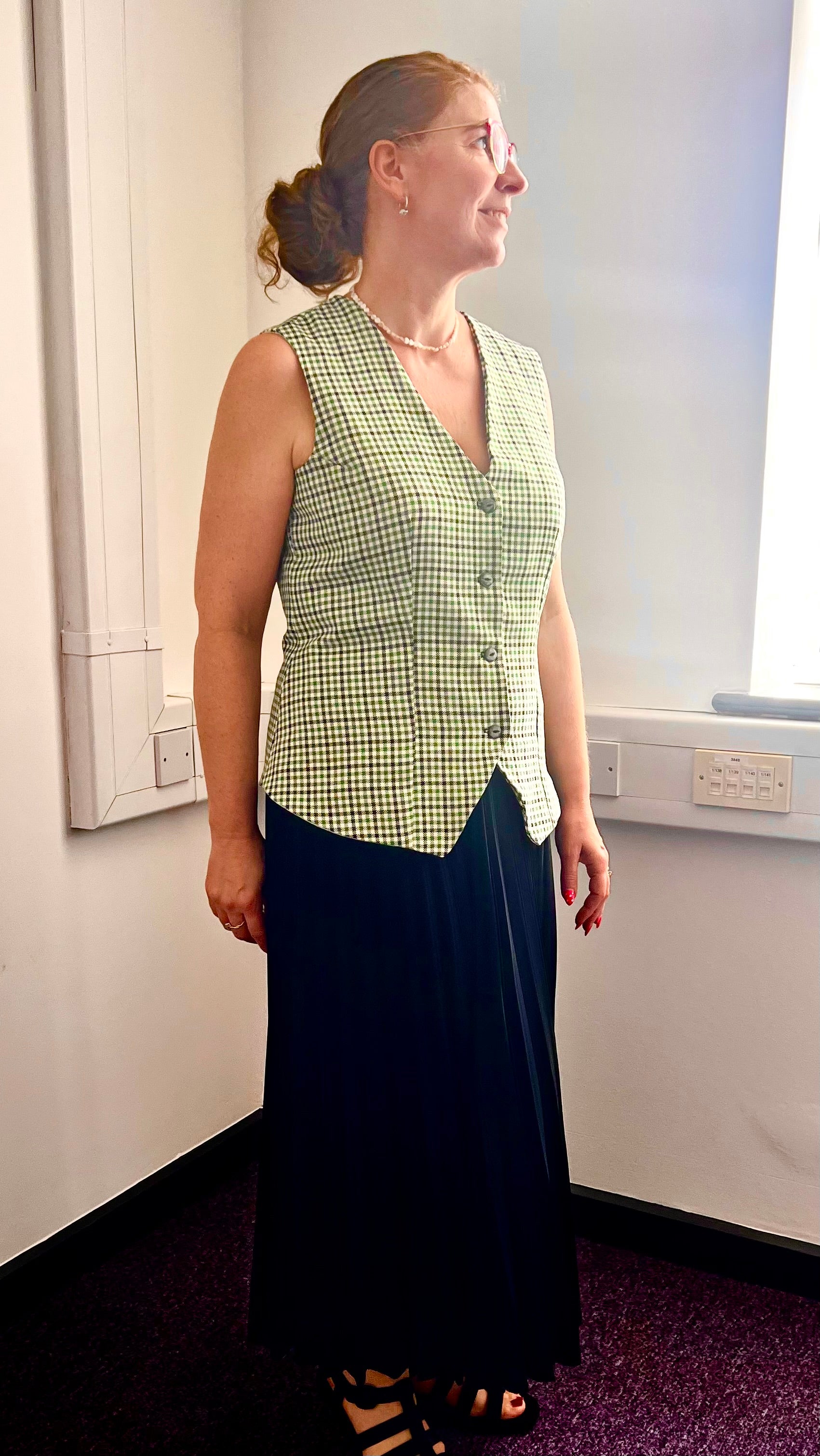 Woman wearing a green checkered vest over a white shirt with a dark skirt indoors.