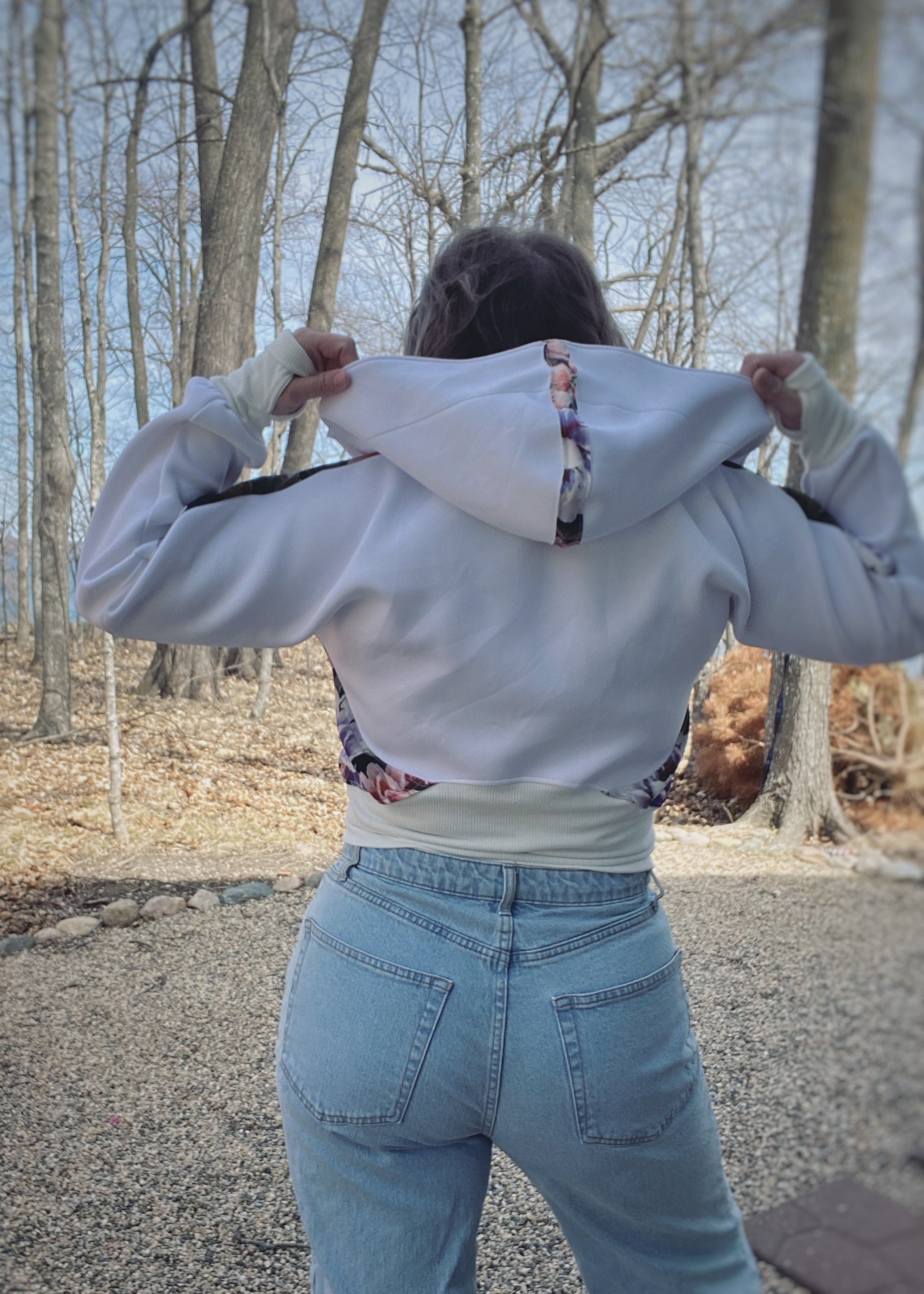 Woman outside by a lake modeling a color-block white and floral athletic bomber jacket with zip closure and scuba hood. Showing the back. Cropped version. Ellie and Mac PDF sewing patterns.