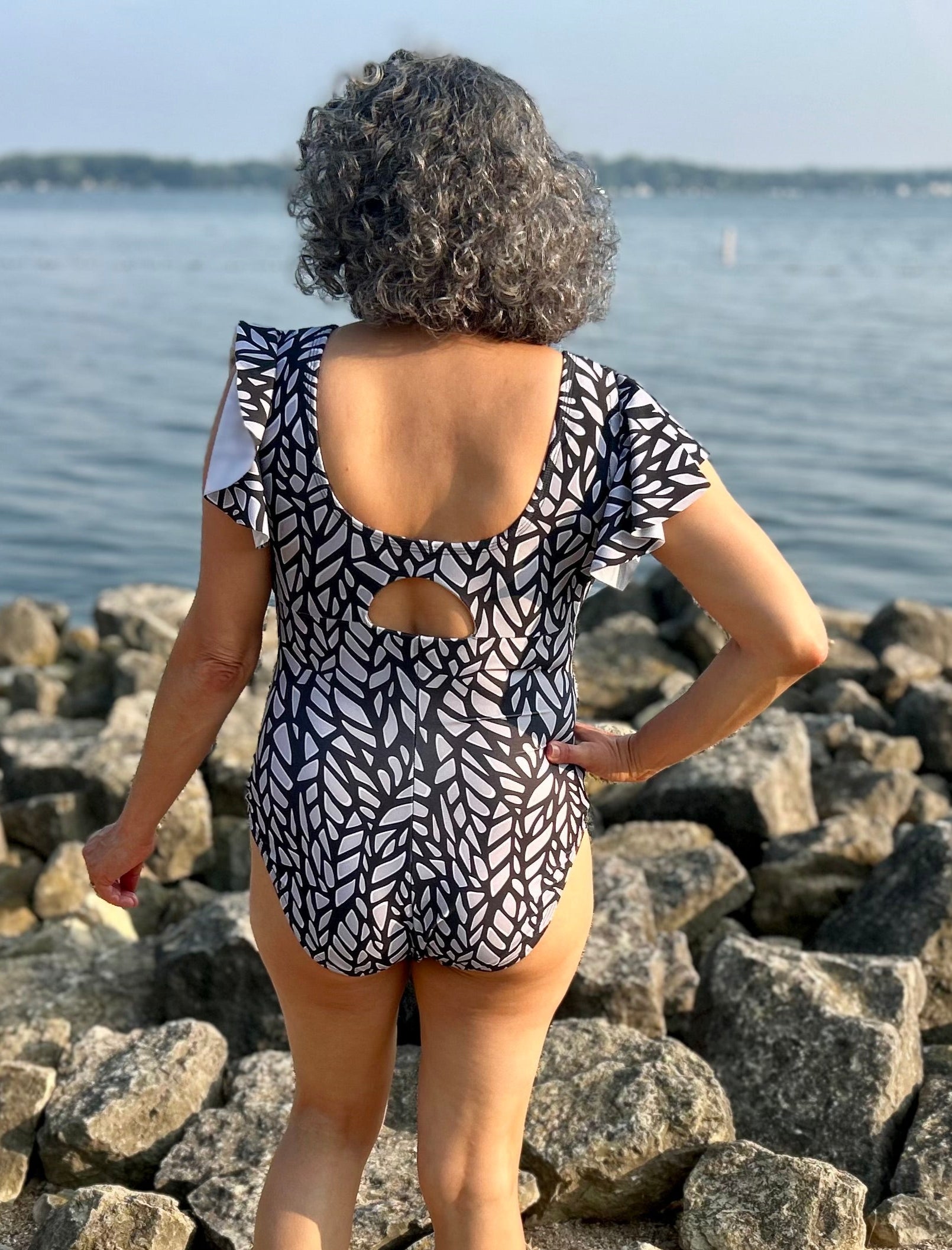 Person wearing a patterned swimsuit standing on rocks by a body of water.