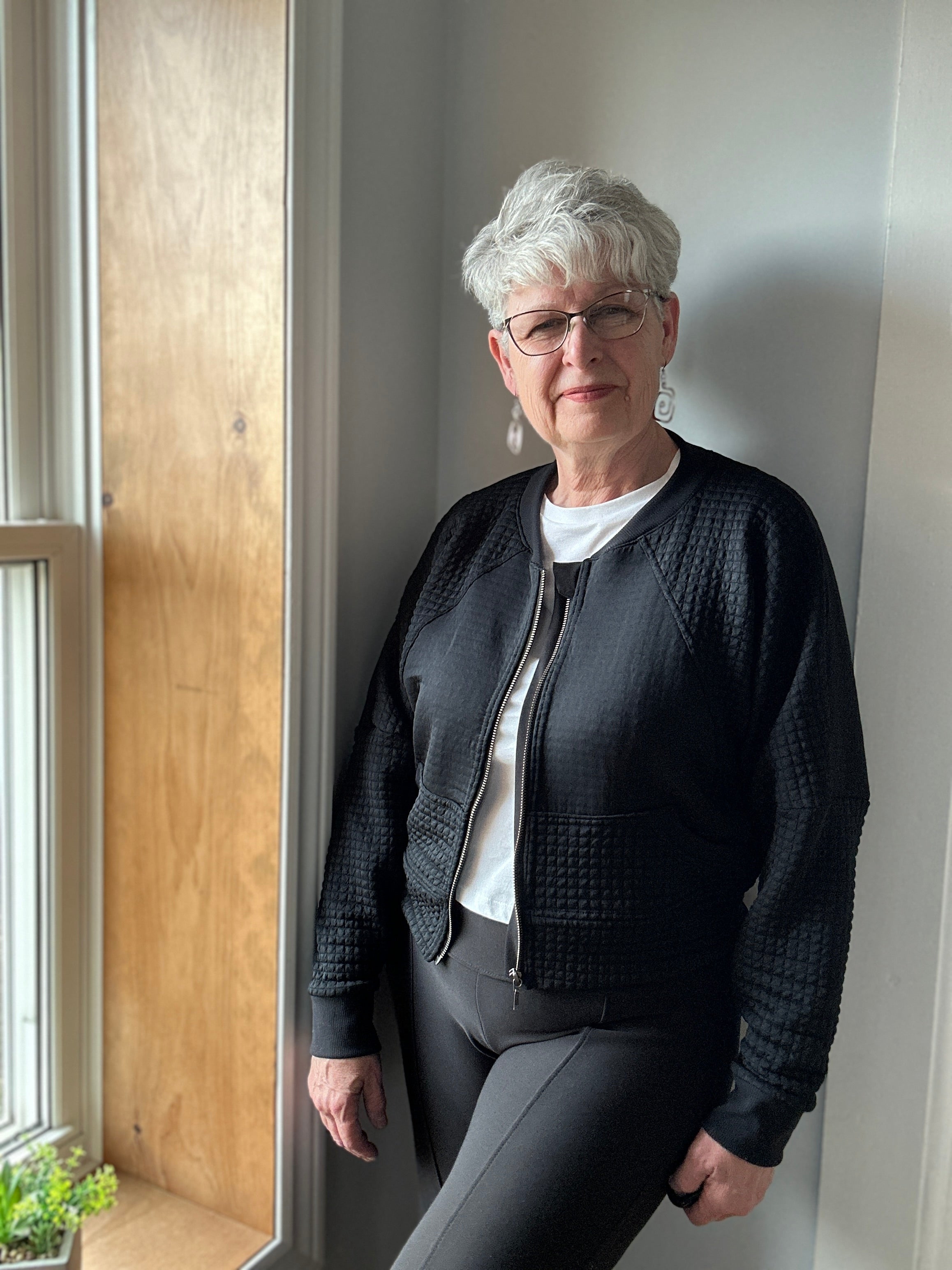 Woman inside by window modeling a black textured fabric knit bomber jacket with zip closure. Athletic Cropped version. Ellie and Mac PDF sewing patterns.