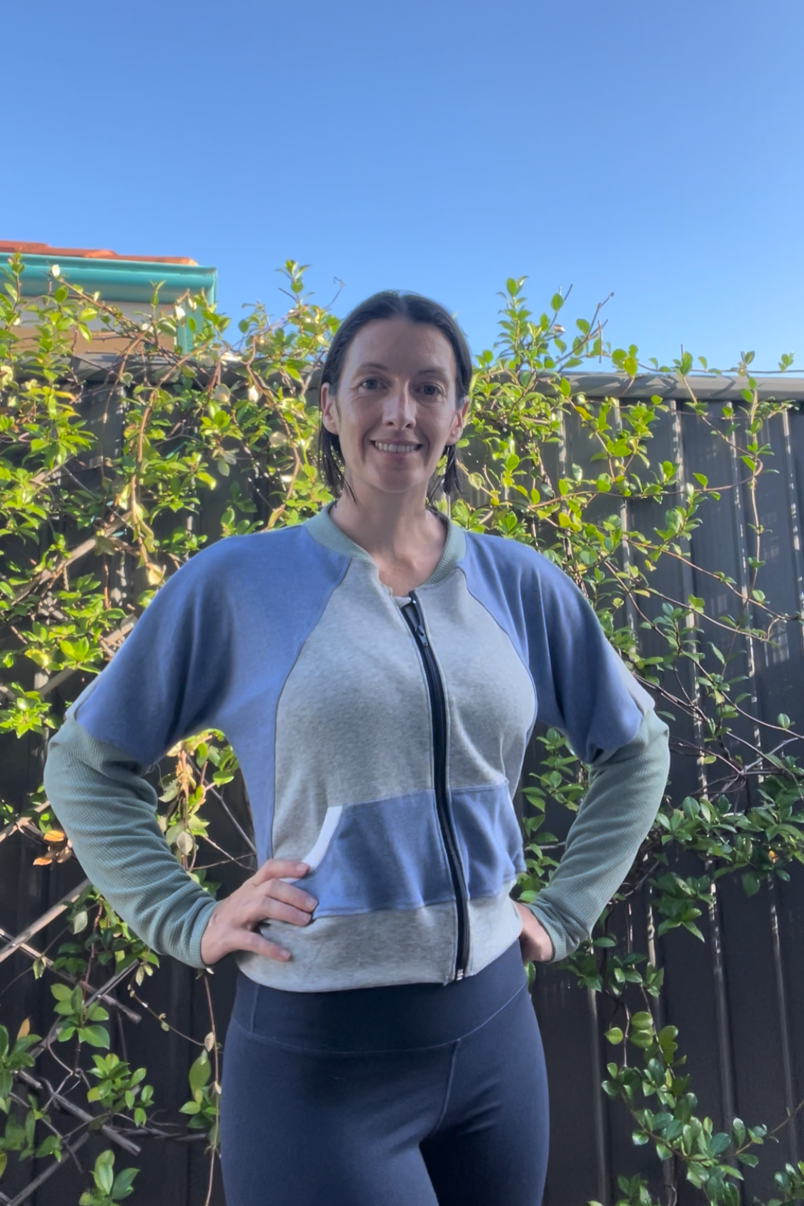 Woman wearing a blue and grey zip-up athletic jacket with raglan dolman sleeves, kangaroo pocket, and curved waistband. Sewing pattern for jackets by Ellie and Mac sewing patterns.