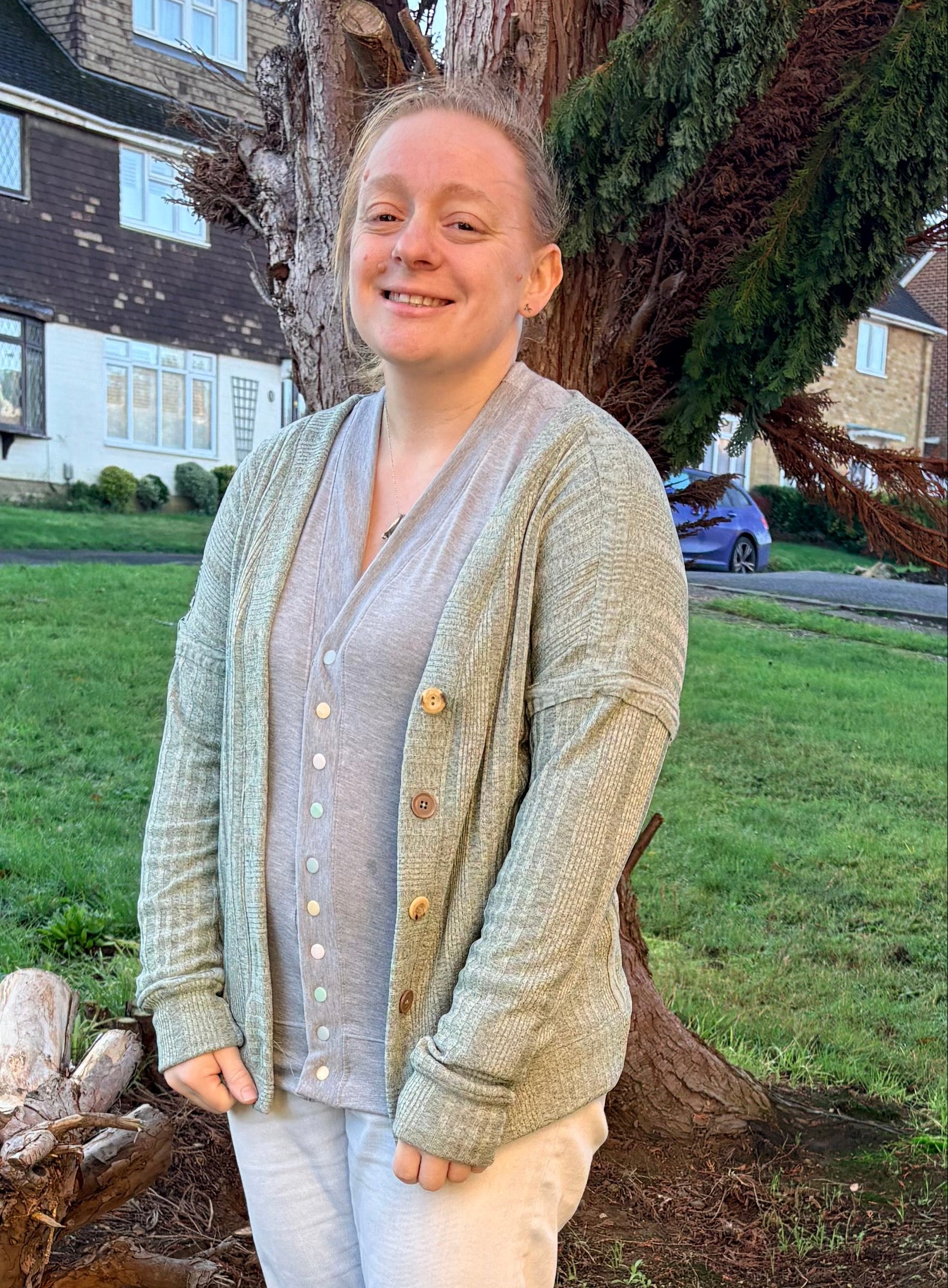 Person standing outdoors in a garden with trees and a house in the background