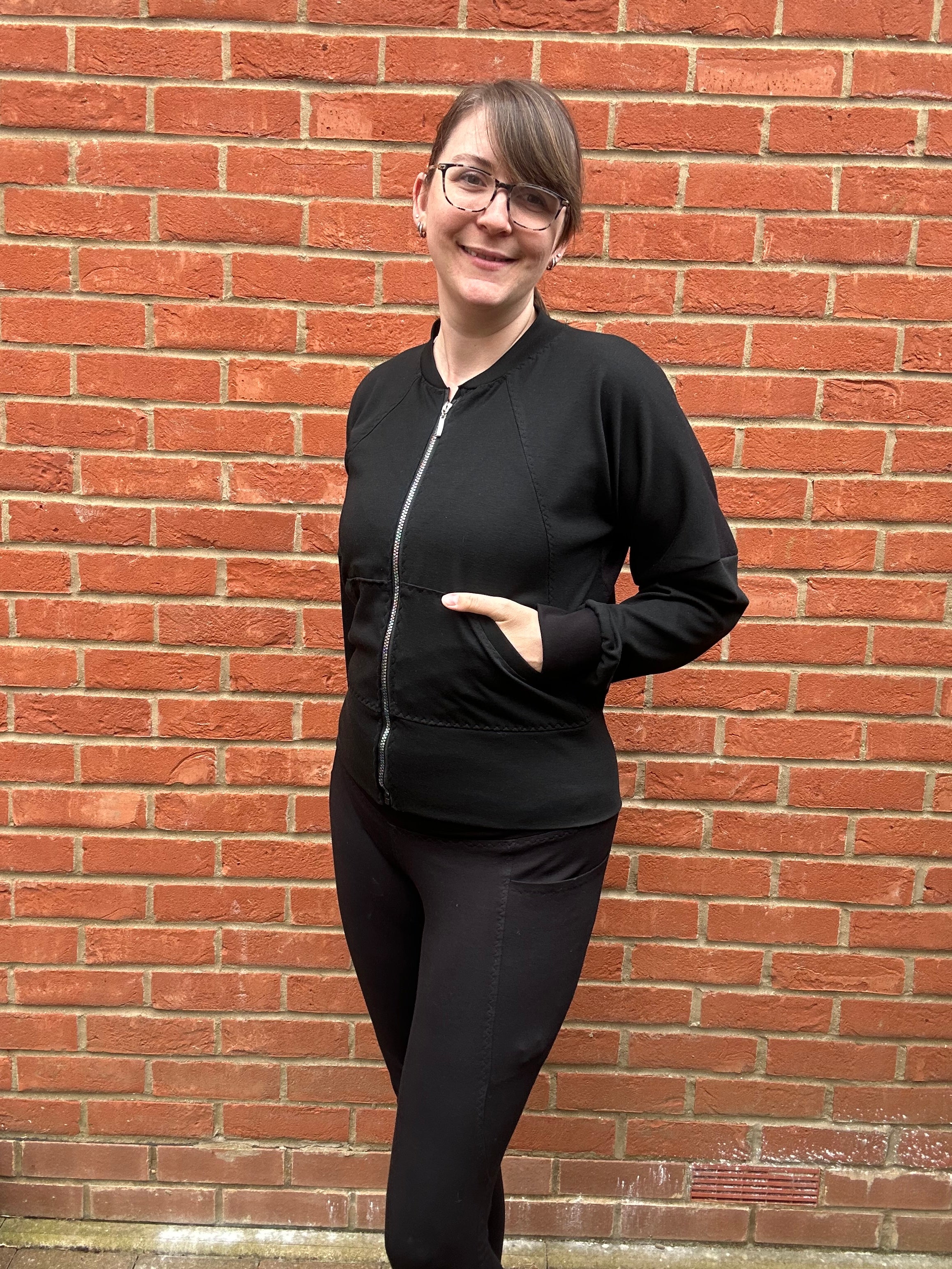 Woman outside by a brick wall modeling a black athletic bomber jacket with zip closure and kangaroo pocket detail. Easy pdf sewing pattern.