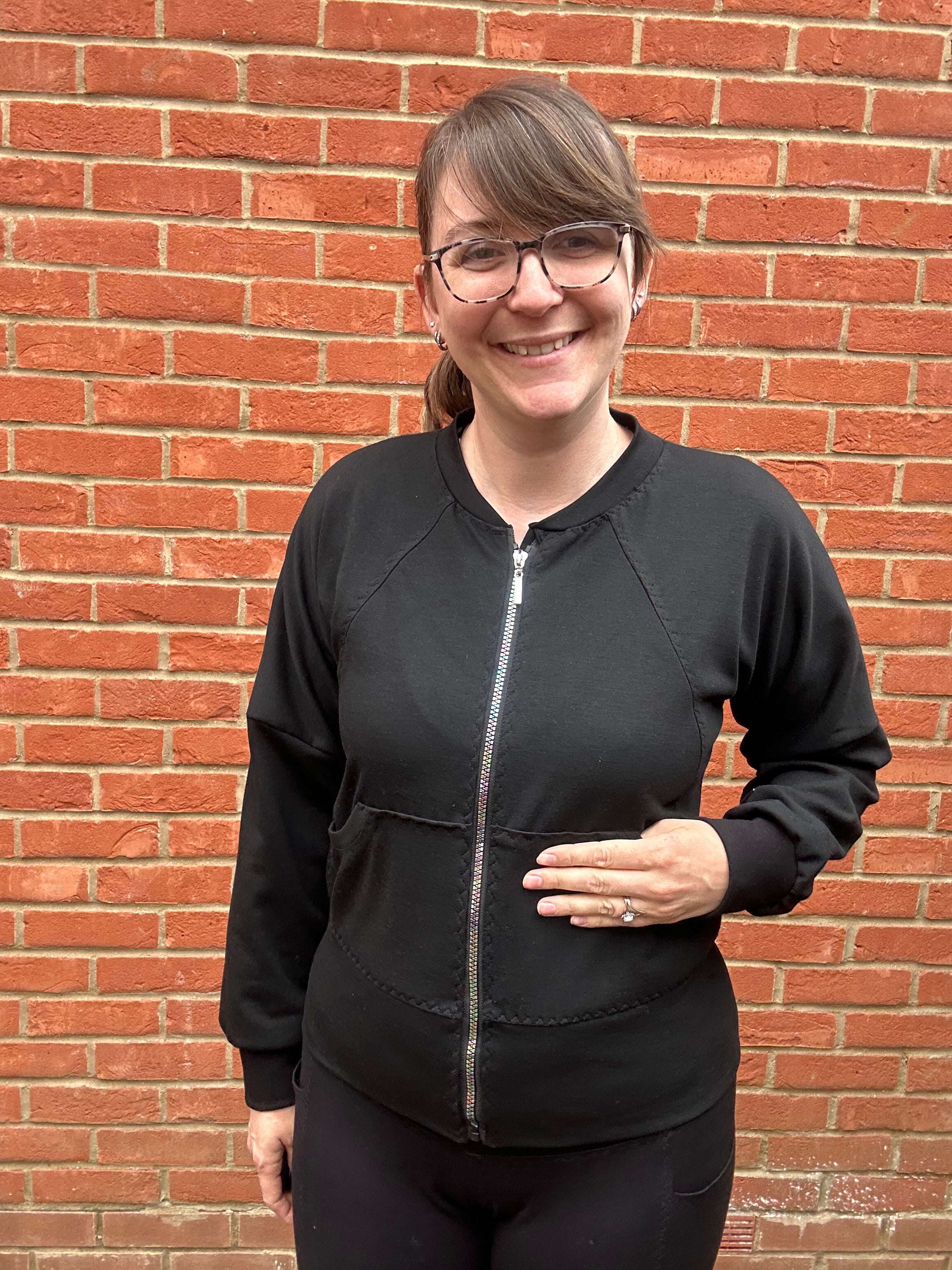 Woman outside by a brick wall modeling a black athletic bomber jacket with zip closure and kangaroo pocket detail. Easy pdf sewing pattern.