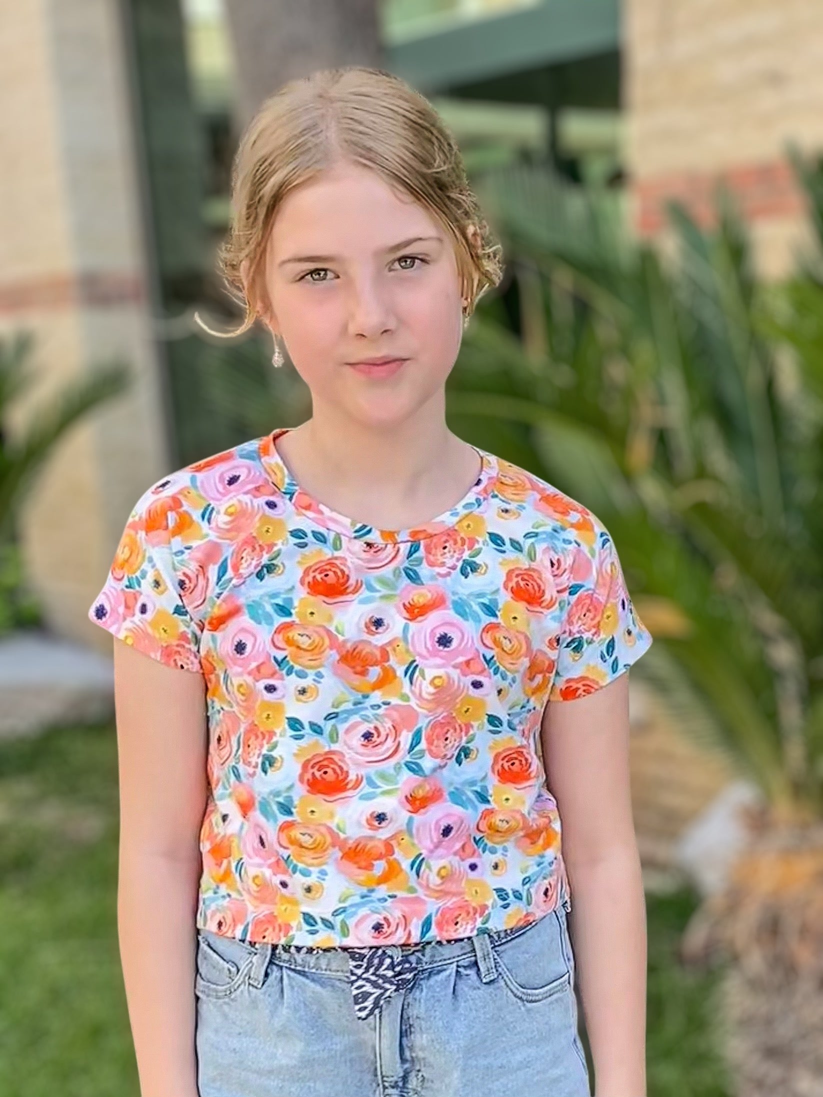 Young girl wearing a colorful floral shirt standing outdoors with plants in the background.