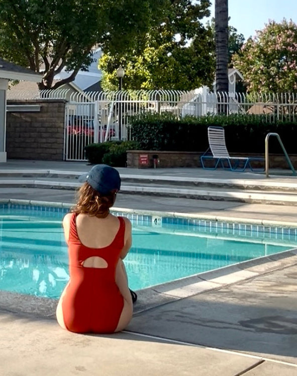 Person in a red swimsuit sitting by a pool with palm trees and a house in the background