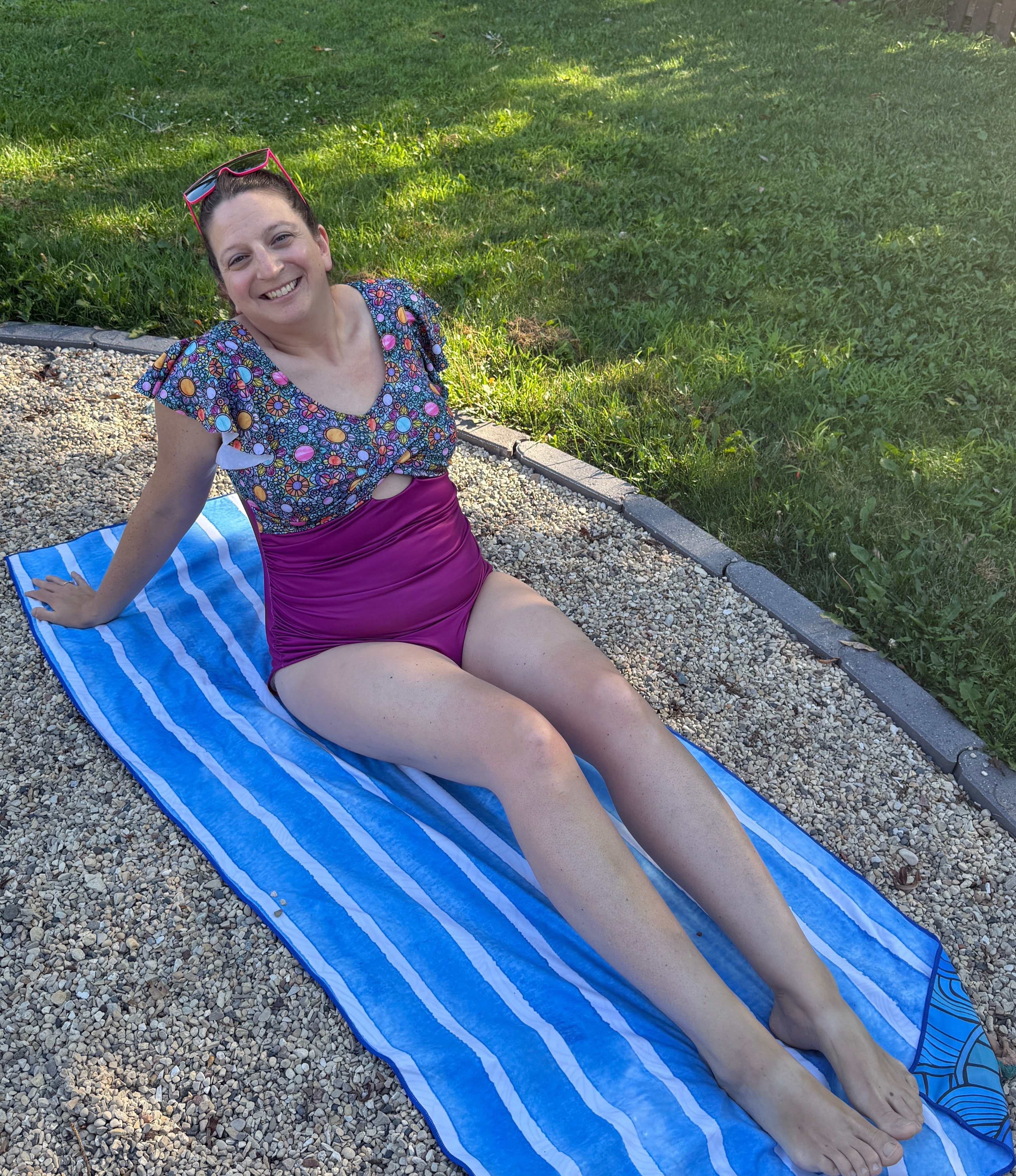 Woman sitting on a blue striped towel in a backyard wearing a burgundy and floral swimsuit. The swimsuit was made with the tropical twist swimsuit pdf sewing pattern. It features cut outs in front and back.