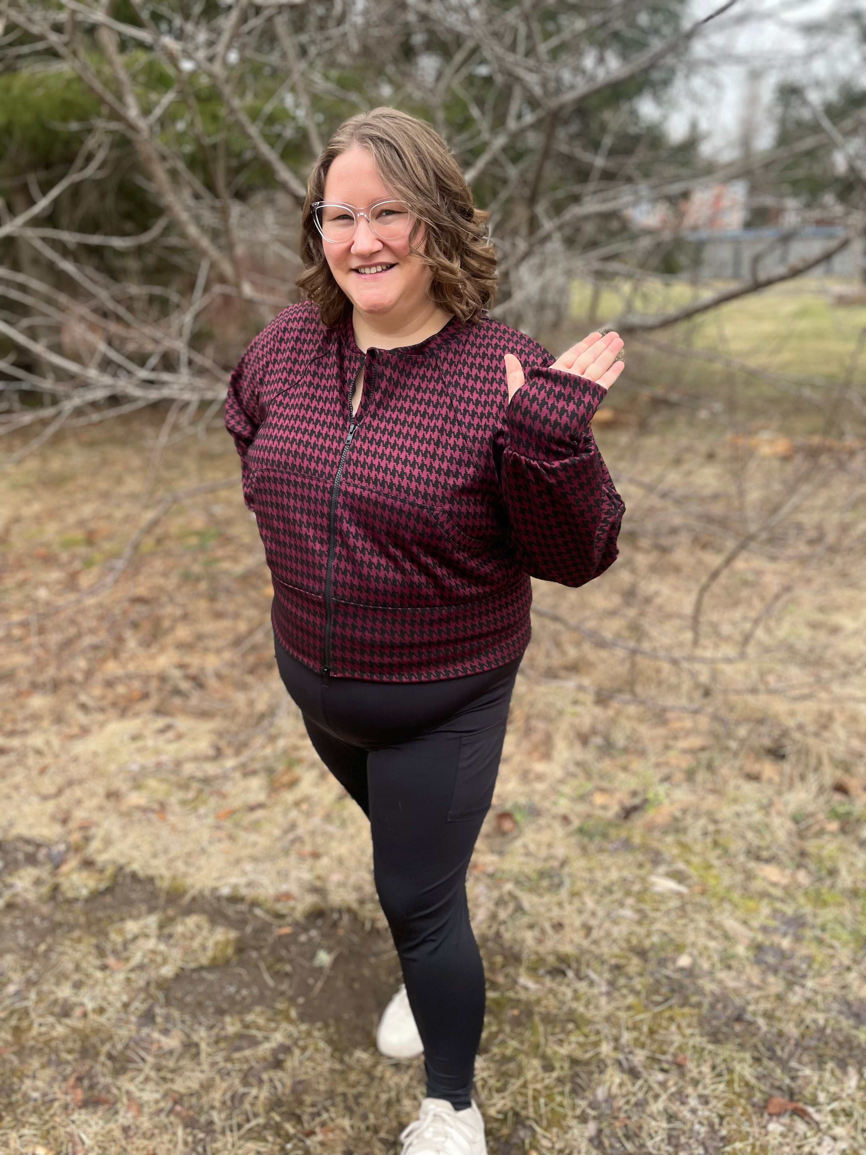 Woman outside modeling a burgundy and black houndstooth athletic bomber jacket with zip closure, kangaroo pocket detail, and thumbhole cuffs. Easy pdf sewing pattern.