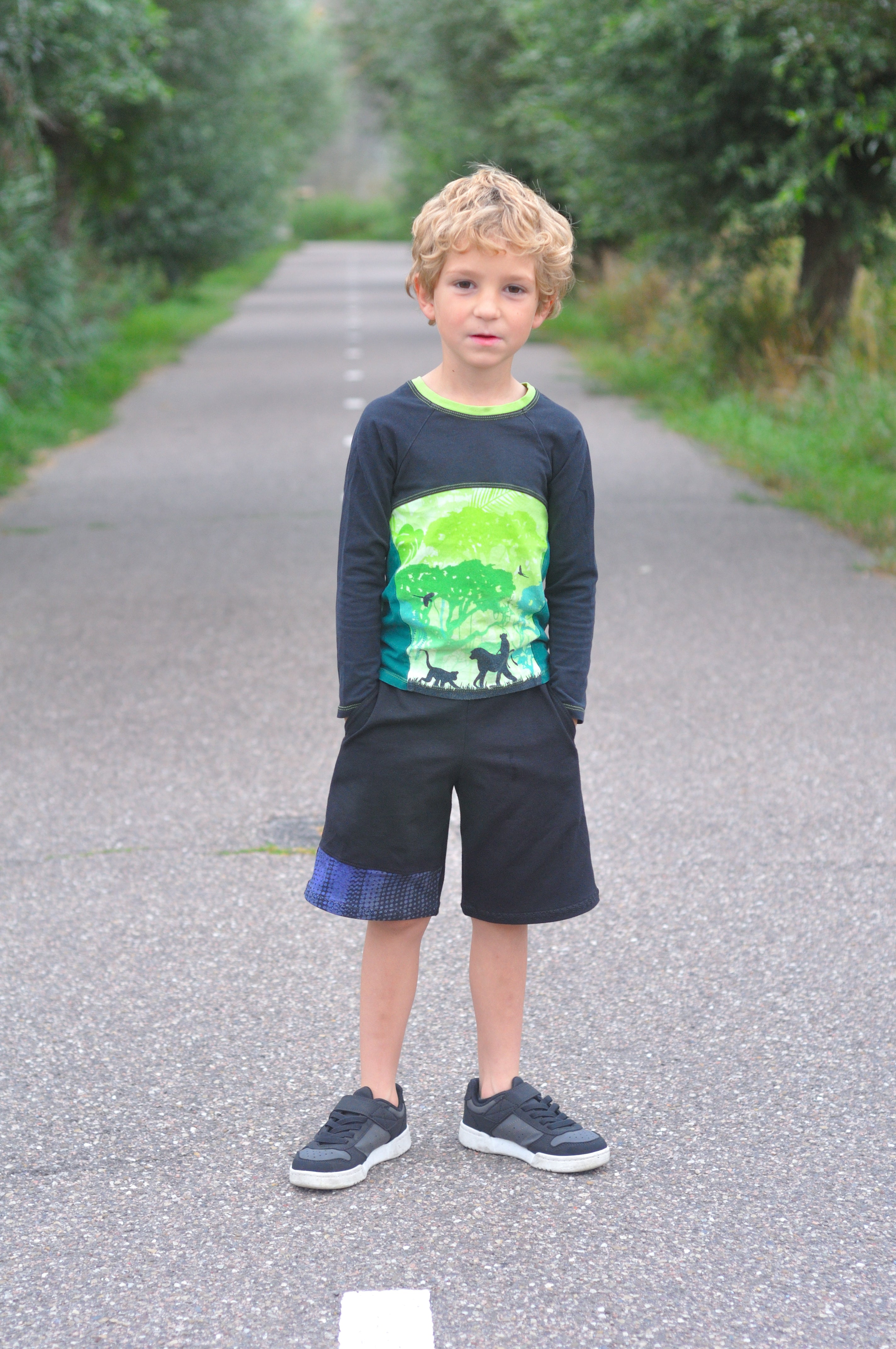 Young boy standing on a path surrounded by greenery wearing a dark blue sweater with a colorful design and black shorts.