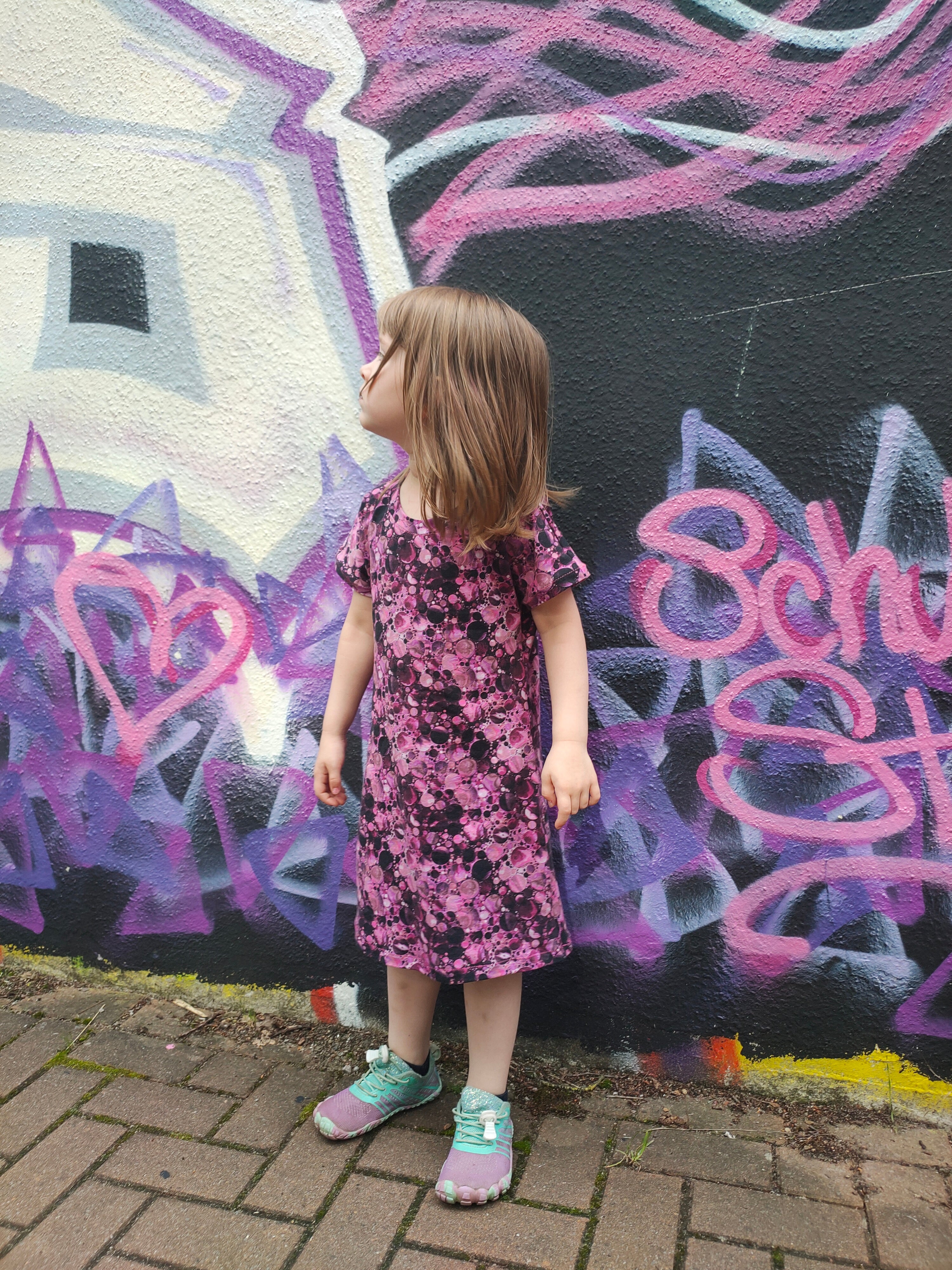 Child in a floral dress standing in front of a colorful graffiti wall. Dress sewing pattern ellie and mac.